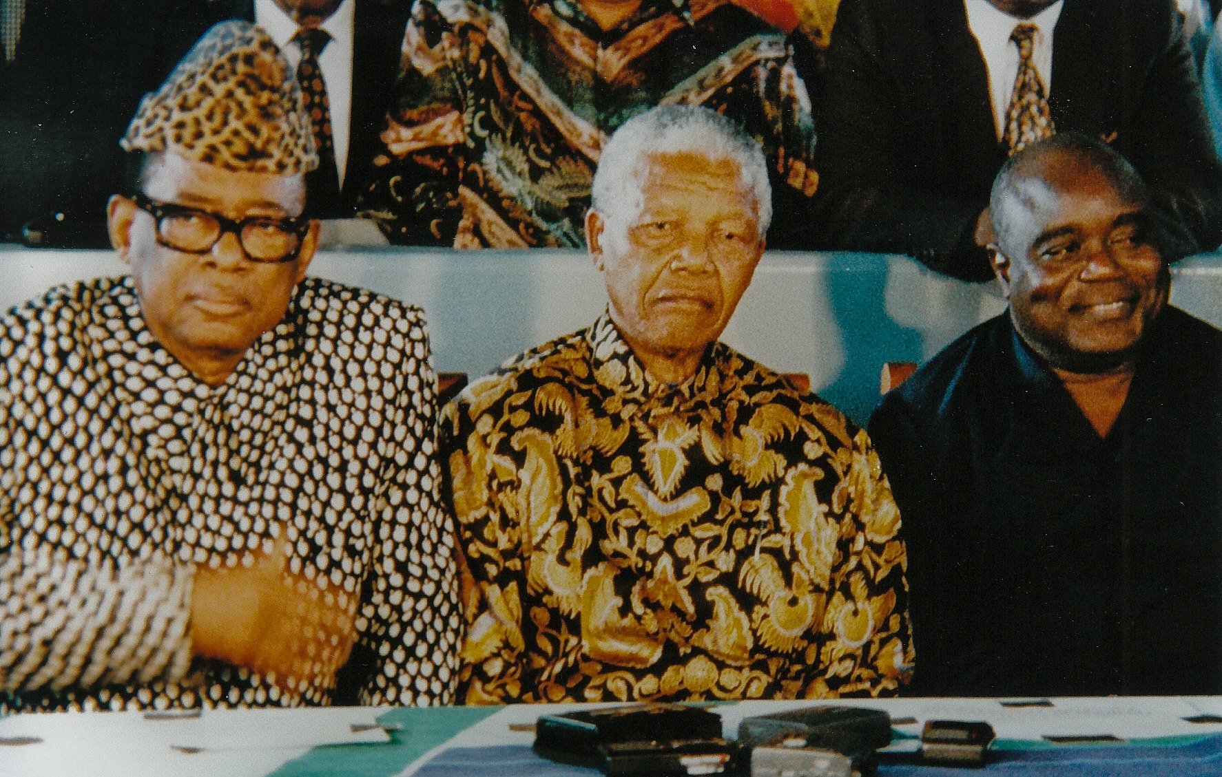 L'image montre trois hommes assis ensemble à une table. Le premier homme, à gauche, porte une chemise ornée d'un motif en damier noir et blanc, ainsi qu'un chapeau avec un motif similaire. Au centre, se trouve un homme célèbre avec une expression sérieuse, vêtu d'une chemise à motifs dorés sur fond noir. À droite, le troisième homme est habillé d'une chemise noire et sourit légèrement. Ils sont tous les trois entourés d'un groupe de personnes en arrière-plan, contribuant à une atmosphère de rassemblement et de discussion. La table devant eux est ornée de divers objets, probablement des appareils d'enregistrement ou des microphones.