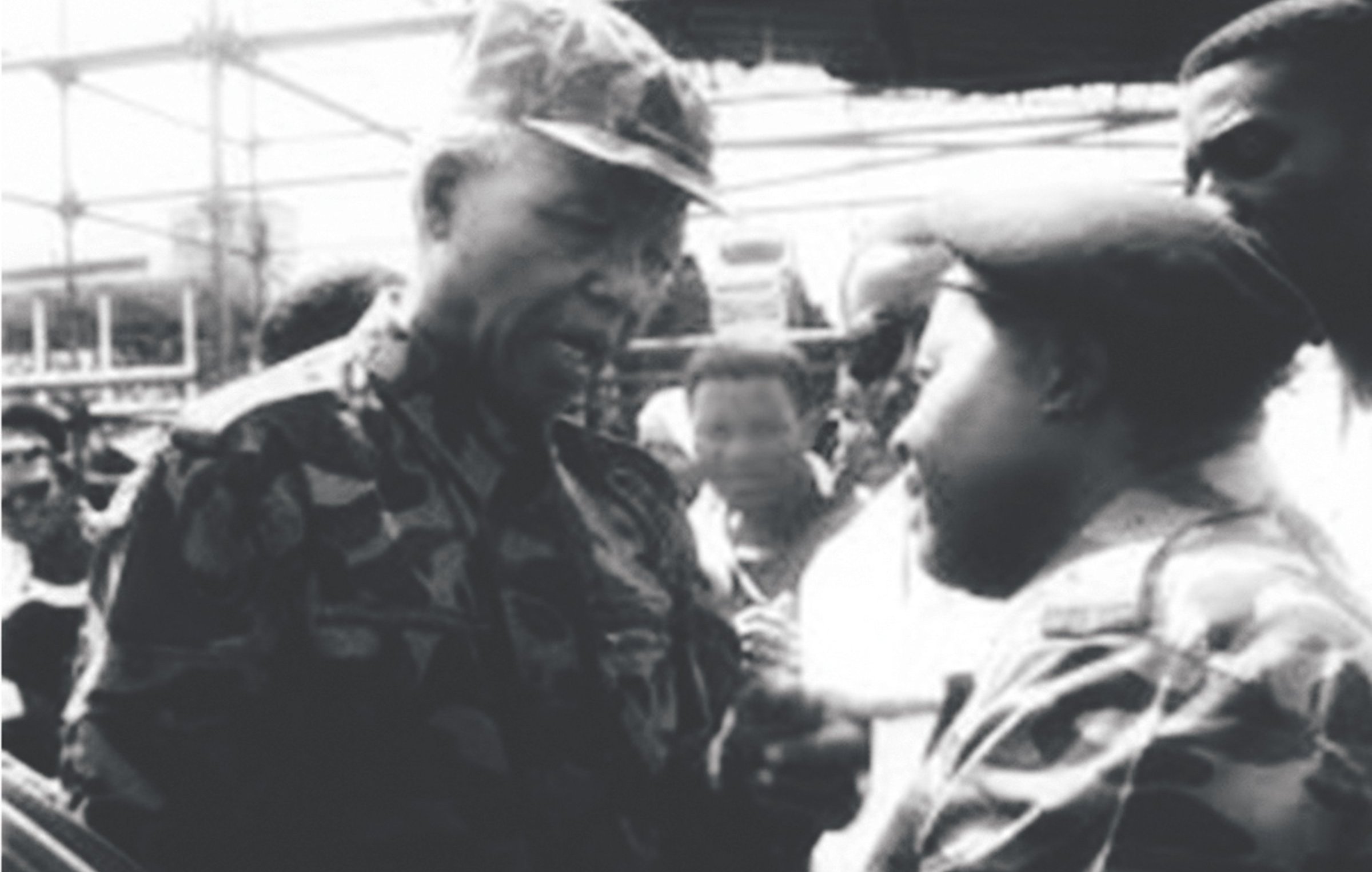 Cette image en noir et blanc montre deux hommes engagés dans une conversation intense. L'homme à gauche, avec un chapeau de camouflage et un uniforme assorti, a une expression sérieuse et attentive. À sa droite, un autre homme portant également une tenue de camouflage semble écouter avec intérêt. En arrière-plan, on peut voir un groupe de personnes, floues, qui regardent la scène. L'atmosphère de l'image suggère un moment significatif, probablement lié à une discussion importante ou à un événement marquant.