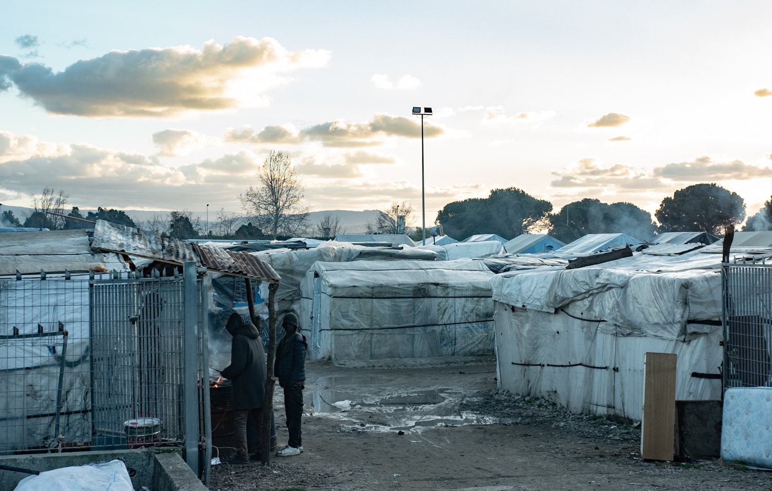 L'image montre un paysage de campement, probablement un bidonville, où des abris temporaires sont installés. Ces structures, couvertes de bâches blanches, sont disposées de manière désordonnée, formant des allées étroites entre elles. Au premier plan, deux personnes se tiennent près d'un feu, suggérant un moment de convivialité ou de chaleur dans un environnement difficile. Le ciel est nuageux, avec des teintes de bleu et d'orange, indiquant une heure de la journée où le soleil se lève ou se couche. En arrière-plan, des arbres et des monts se dessinent, ajoutant une dimension naturelle à ce paysage urbain. L'atmosphère peut sembler mélancolique, mais aussi résiliente, avec des traces de vie et d'activités humaines.