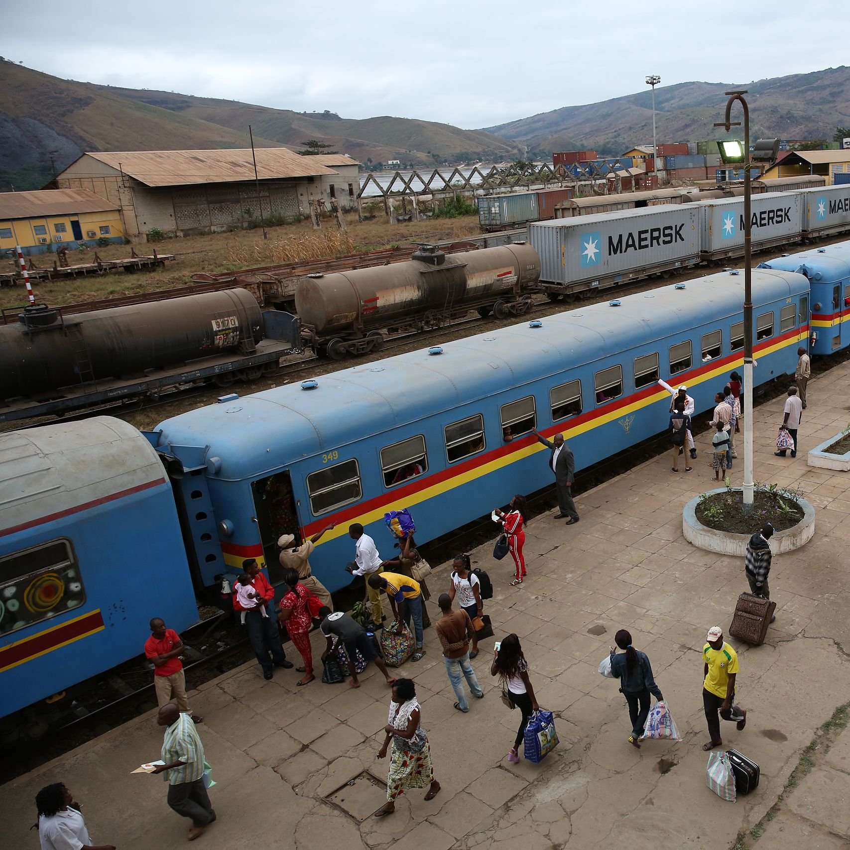 L'image montre une gare animée. De longs trains de marchandises et de passagers sont alignés sur les quais. Les wagons sont d'un bleu vif avec des bandes colorées. Des voyageurs descendent et montent à bord, portant des sacs et des effets personnels. On peut entendre des conversations, des annonces de train, et le bruit des roues sur les rails. En arrière-plan, on aperçoit des collines qui entourent la gare. L'atmosphère est vive et dynamique, avec un mélange de mouvements et d'interactions entre les passagers.