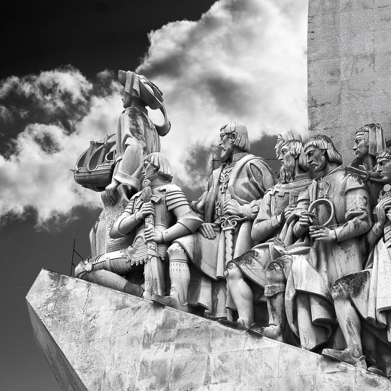 L'image représente un relief sculpté monumentale. On y voit une procession de figures humaines, probablement des explorateurs ou des conquérants, qui se tiennent debout sur une plateforme. Les personnages sont vêtus de costumes historiques et affichent des expressions déterminées. Certains d'entre eux portent des armes, des drapeaux et d'autres objets symboliques, témoignant de leurs exploits. En arrière-plan, on aperçoit des nuages sombres qui ajoutent une atmosphère dramatique à la scène. Le tout est sculpté dans un matériau clair, probablement de la pierre, permettant de bien contraster avec le ciel nuageux. Cette œuvre dégage une impression de puissance et de grandeur, évoquant une célébration de l'histoire et de l'aventure.