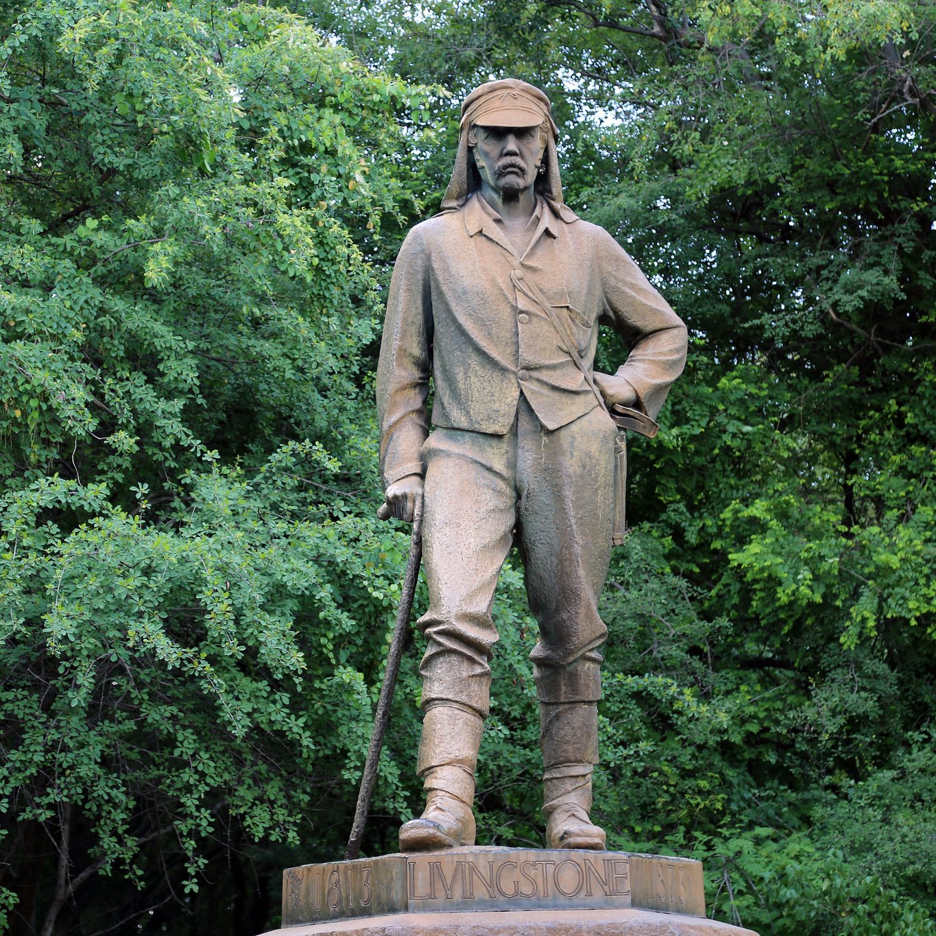 L'image montre une statue en bronze d'un homme qui se tient debout, avec une posture confiante. Il porte des vêtements de style explorateur du XIXe siècle, incluant une chemise à manches longues et un pantalon. Son regard est déterminé et il a une canne à la main, suggérant qu'il pourrait être un aventurier ou un explorateur. La statue est placée sur un socle avec une inscription, et elle est entourée d'une végétation dense, avec des arbres verts en arrière-plan, créant un contraste entre la statue et la nature environnante. L'atmosphère est calme et sereine, reflétant peut-être le respect pour cette figure historique.