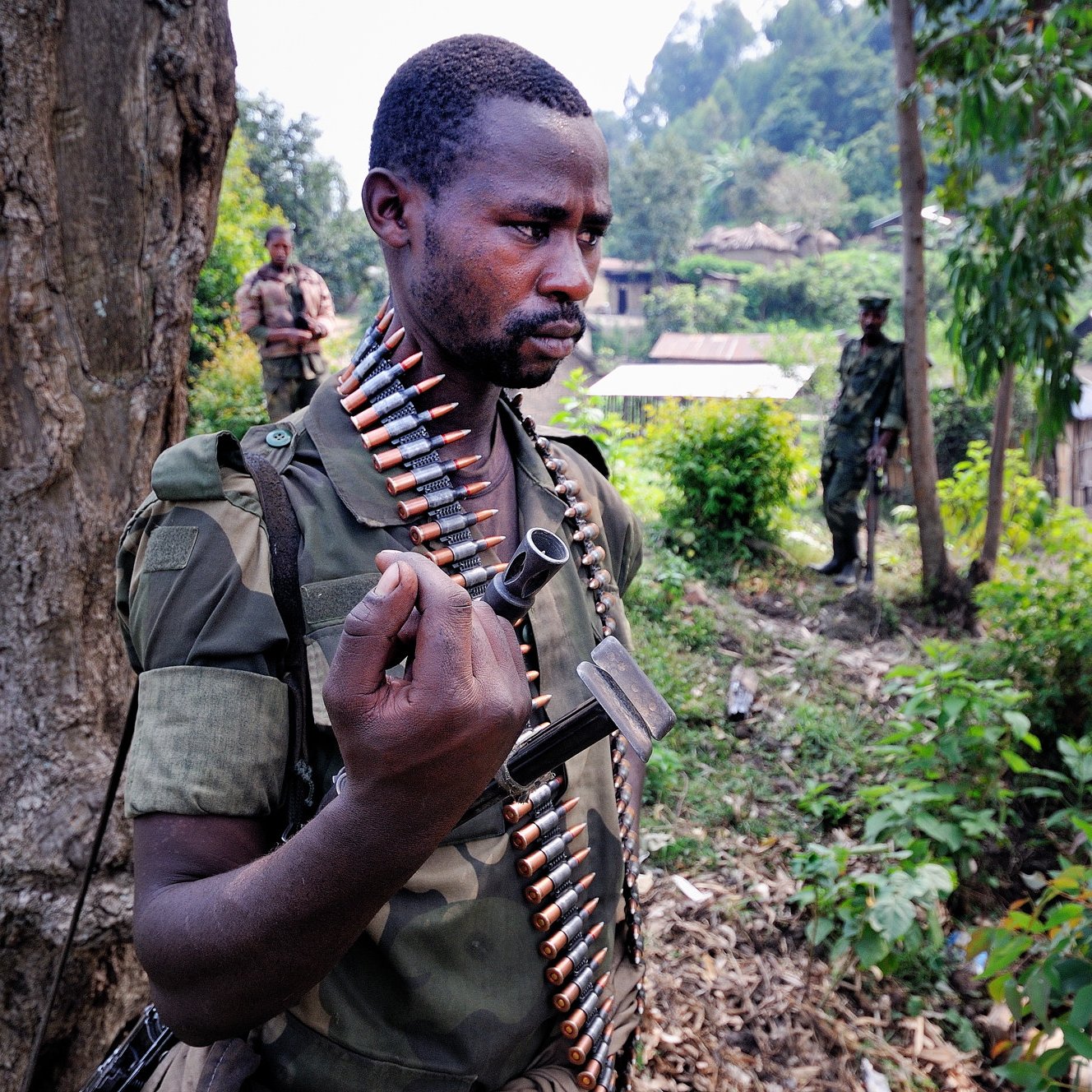 L'image montre un soldat debout près d'un arbre, portant un uniforme militaire camouflage. Il a une ceinture de munitions autour de son torse, et il tient une arme à la main. Son expression est sérieuse, indiquant la concentration et la vigilance. En arrière-plan, d'autres soldats sont visibles, et le paysage est verdoyant, avec des habitations simples au loin. L'atmosphère dégage une impression de tension en raison du contexte militaire.