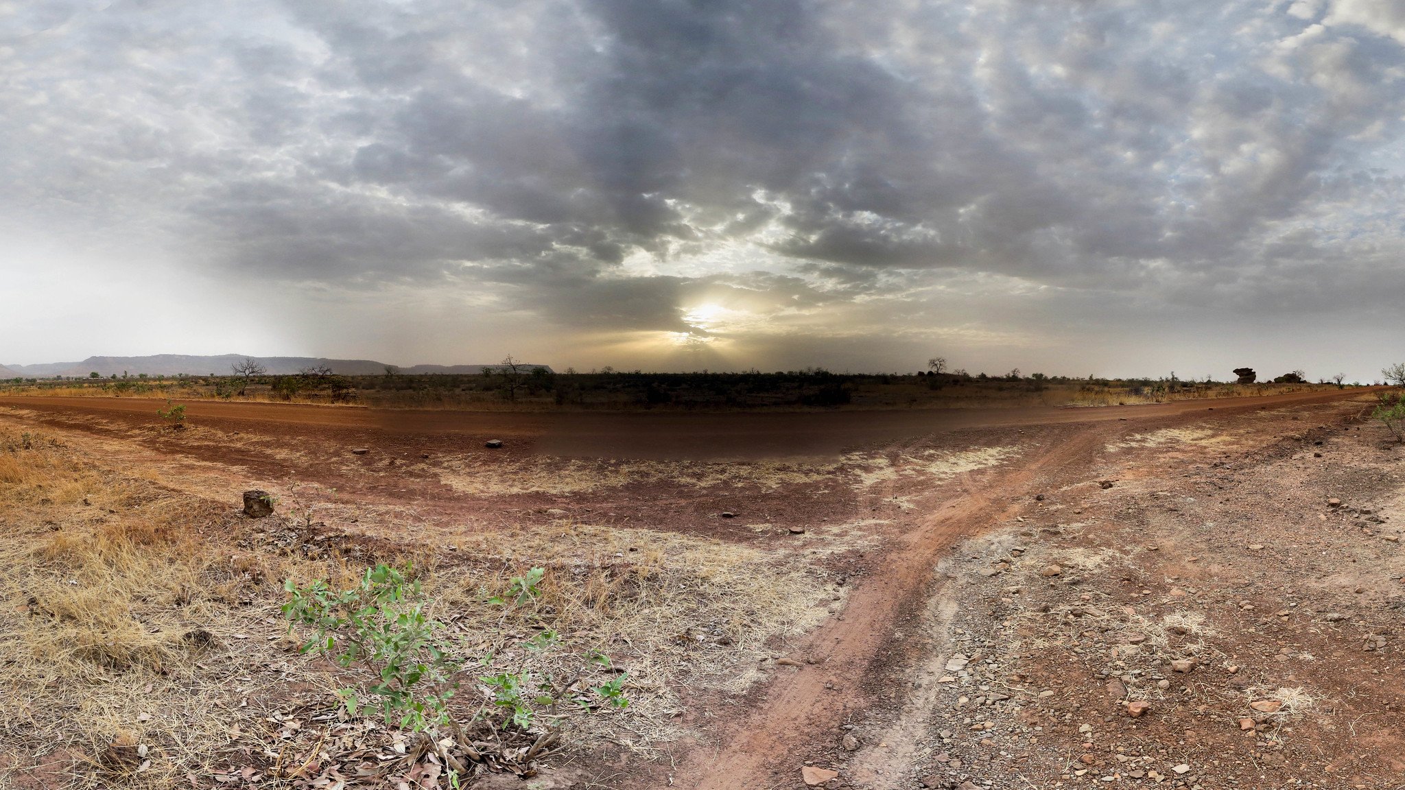 Imaginez un vaste paysage ouvert, dominé par des tons chauds de terre et des nuances de beige. Le sol est sec et parsemé de petites pierres, avec des herbes jaunies qui émergent de la terre aride. À l'horizon, un ciel nuageux s'étend, teinté de gris avec des rayons de lumière qui percent, annonçant un coucher de soleil. La lumière crépusculaire crée une ambiance paisible, tandis que la nature environnante semble calme et tranquille. Les silhouettes lointaines de collines ajoutent une dimension au paysage, renforçant le sentiment d'immensité et d'isolation.