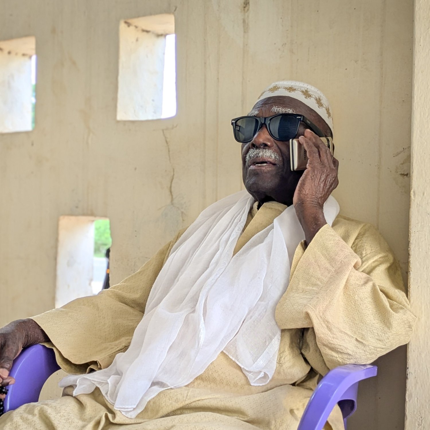 L'image montre un homme assis sur une chaise en plastique violette, contre un mur de couleur beige avec des fenêtres rectangulaires qui laissent passer une lumière douce. Il porte des lunettes de soleil noires et un vêtement traditionnel de couleur claire, accompagné d'un châle blanc drapé sur ses épaules. L'homme est en train de parler au téléphone, son visage exprime la concentration et la sérénité. L'environnement est calme, évoquant un moment de détente dans un cadre paisible.