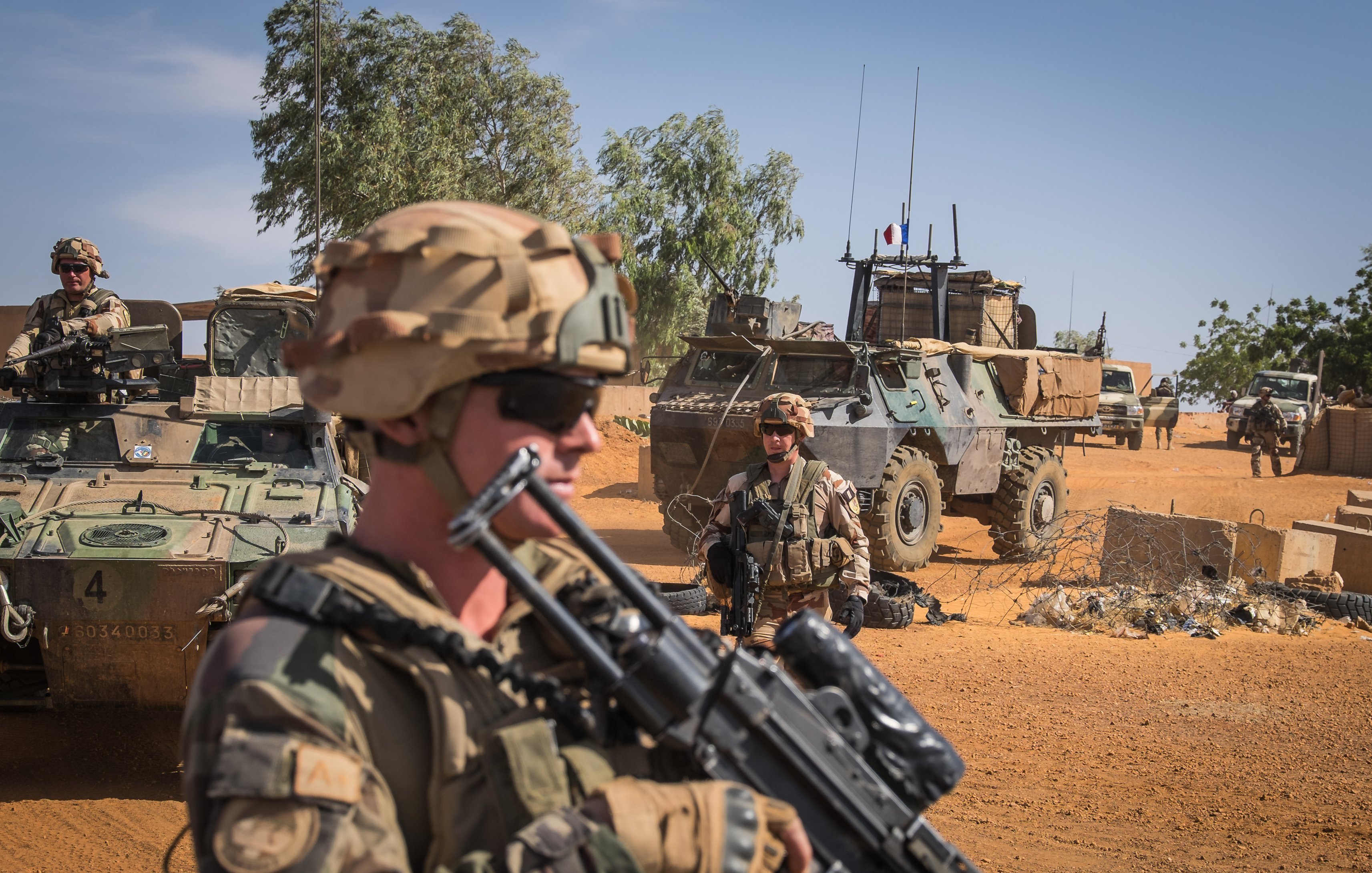 The image depicts a military scene in a desert environment. In the foreground, a soldier wearing a helmet and sunglasses is holding an assault rifle, looking serious and vigilant. Behind him, there are other soldiers and military vehicles, including an armored personnel carrier. The background shows a clear blue sky with some greenery and sand, indicating a remote and arid location. The overall atmosphere suggests a sense of readiness and military operations.
