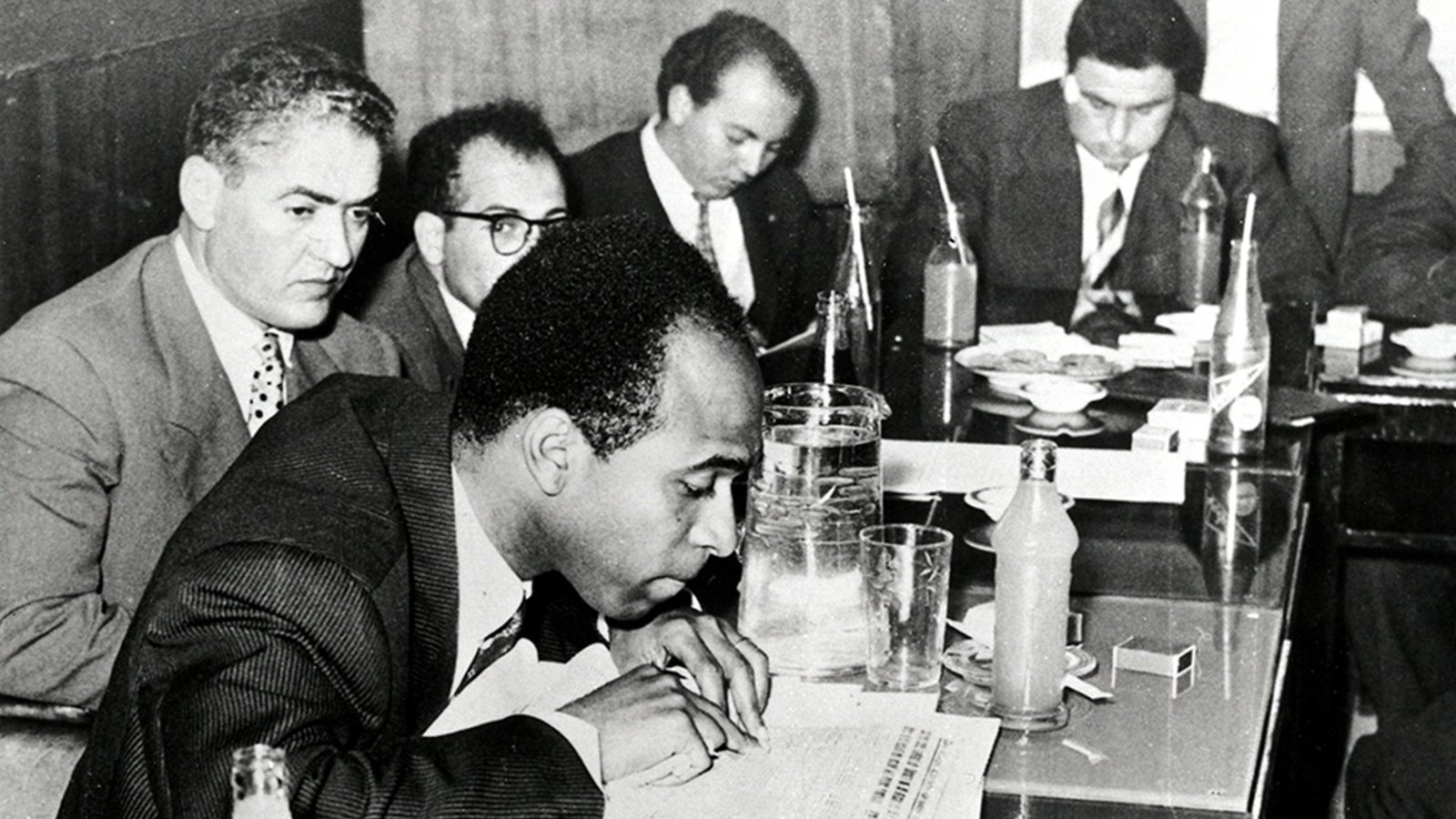 L'image en noir et blanc montre une scène de réunion dans un environnement intérieur, probablement une salle de conférence. Plusieurs hommes sont assis autour d'une table. Au premier plan, un homme est penché sur une feuille de papier, prenant semble-t-il des notes. Il est vêtu d'un costume sombre et d'une cravate, et semble concentré. À sa droite, un autre homme regarde vers le bas, tandis que les autres hommes en arrière-plan discutent ou examinent des documents. Sur la table, on peut voir des verres, des bouteilles, et quelques objets éparpillés. L'atmosphère semble sérieuse et studieuse, évoquant un moment important de collaboration ou de décision.