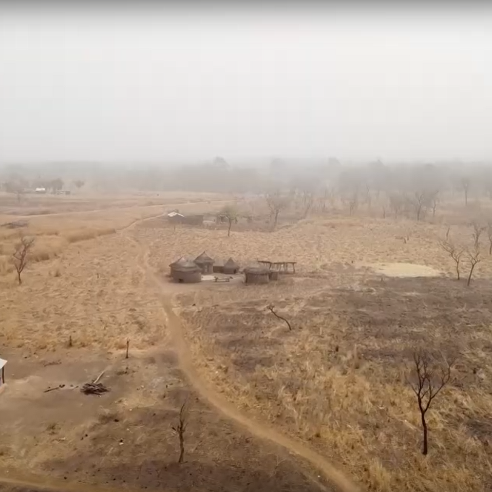 The image depicts a rural landscape characterized by dry, barren land. There are patches of brown grass and sparse vegetation, indicating an arid environment. In the foreground, a small structure with a white roof sits alongside a dirt path. In the background, a few more buildings can be seen, scattered amidst the landscape. The atmosphere appears hazy, possibly due to dust or smoke, giving the scene a muted and subdued appearance. Sparse trees dot the area, and the overall setting suggests a remote, pastoral region.