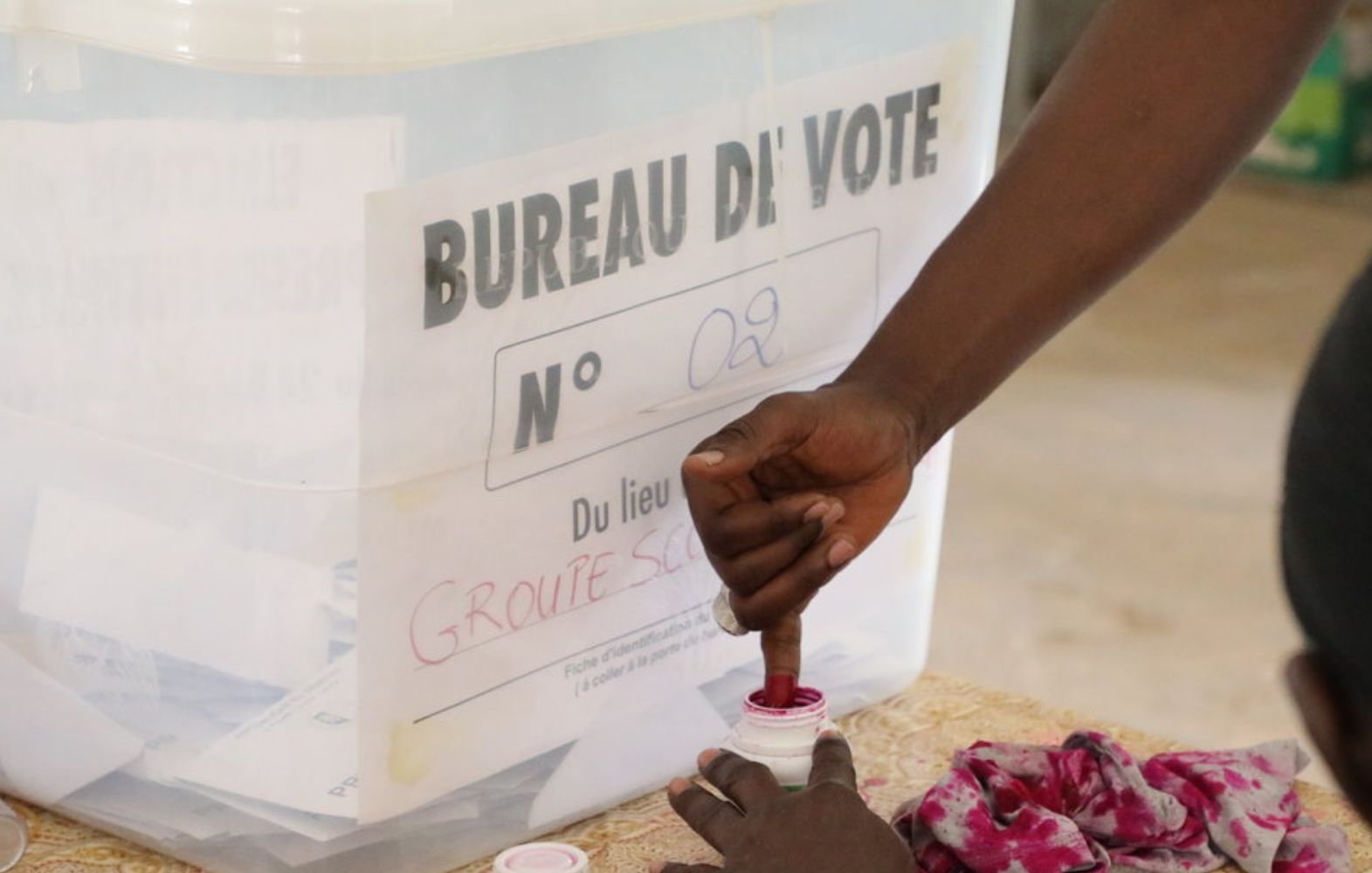 Devant vous se trouve une scène typique d'un bureau de vote. Il y a une urne transparente avec une étiquette indiquant "BUREAU DE VOTE N° 02" et le lieu sur une feuille, probablement fixée à l'urne. Un électeur est en train de voter : sa main est penchée vers une petite boîte de vote où il semble plonger son doigt dans de l'encre rouge. Sur la table, il y a des morceaux de tissu, probablement utilisés pour marquer les bulletins ou pour l'organisation du vote. L'ambiance est calme, avec un sentiment de sérieux et d'engagement civique.