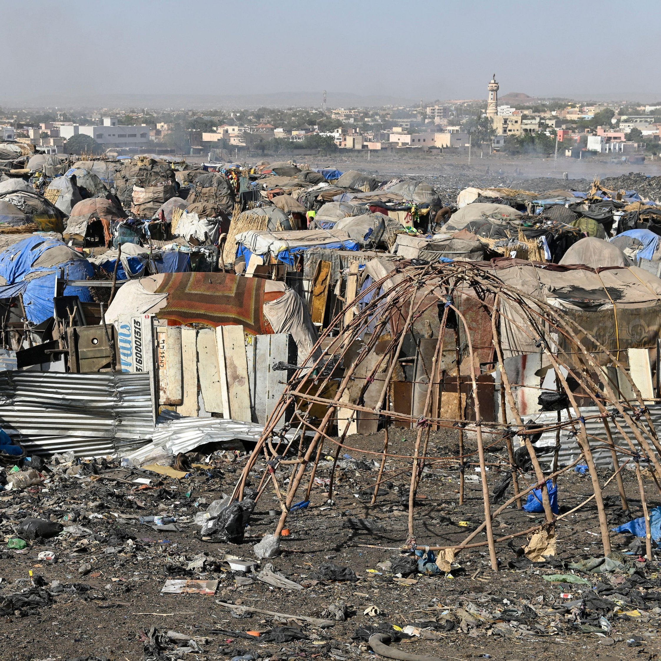 L'image montre un paysage de bidonville, où l'on peut distinguer des habitations précaires construites à partir de matériaux recyclés tels que des tôles, des bâches en plastique et des branches de bois. Ces structures sont souvent assez rudimentaires et forment des petites communautés. Le sol est jonché de déchets et de débris divers, reflétant une forte pollution. À l'arrière-plan, on aperçoit des bâtiments en dur, ainsi qu'une mosquée avec son minaret, indiquant la présence d'une ville proche. L'atmosphère dégage une impression de désolation, mais aussi de vie, avec des traces humaines visibles malgré les conditions difficiles.