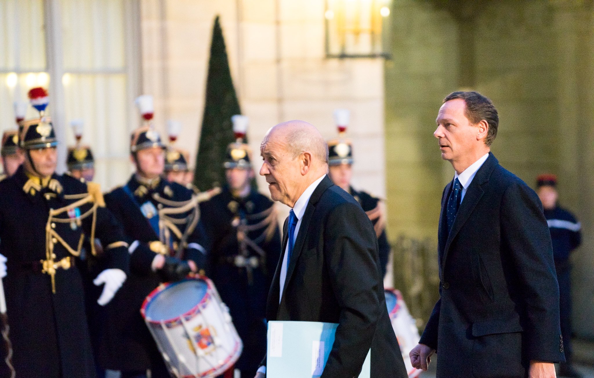 L'image montre deux hommes marchant dans une ambiance officielle. À l'arrière-plan, on peut apercevoir une garde cérémonielle, avec des soldats en uniforme traditionnel, portant des chapeaux distinctifs et tenant des instruments de musique, comme des tambours. Les hommes au premier plan semblent se rendre à une réunion importante, l'un d'eux portant un dossier ou un document. L'éclairage de la scène est doux, créant une atmosphère solennelle et respectueuse.