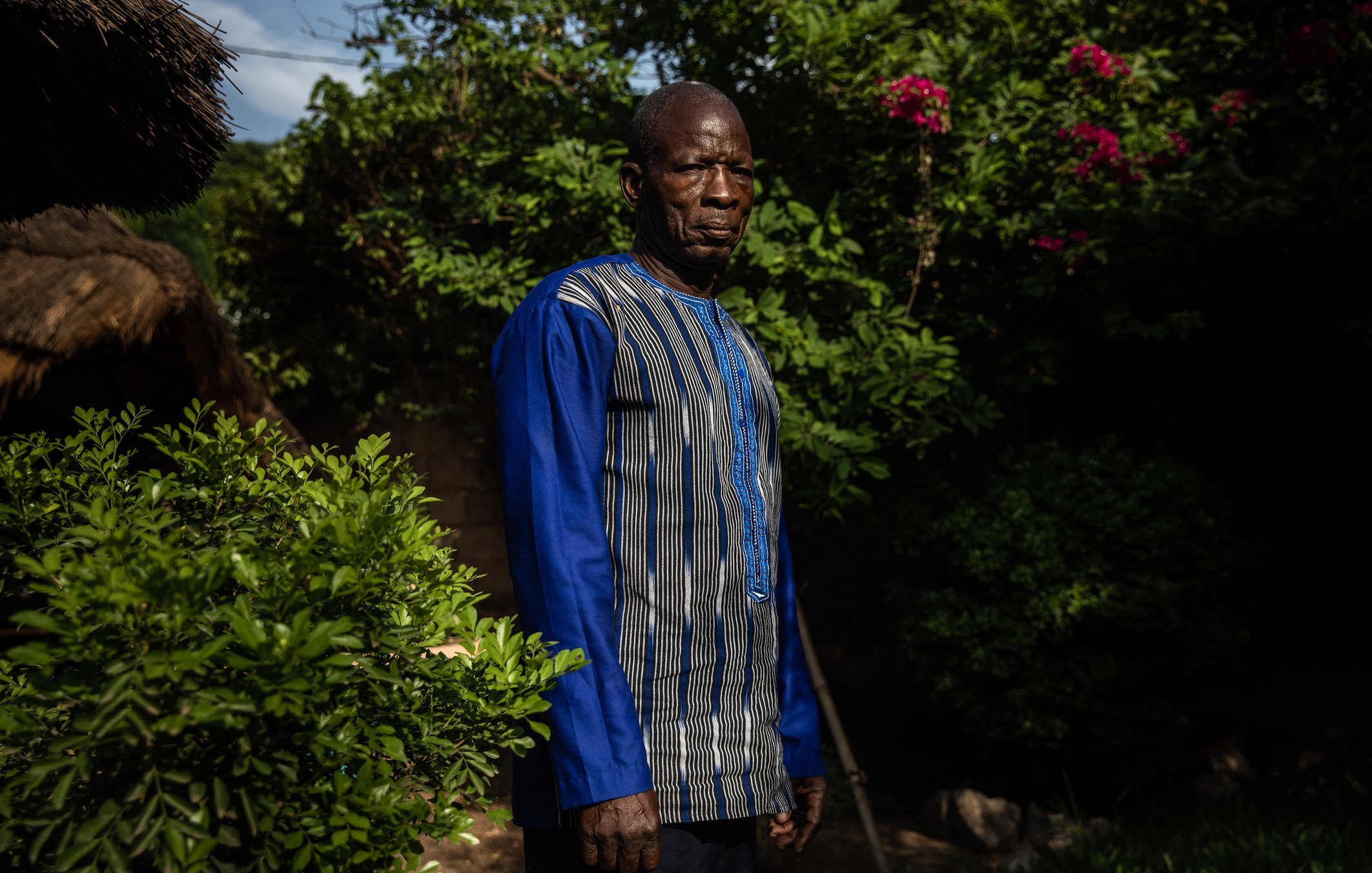 L'image montre un homme debout dans un environnement verdoyant. Il porte une tunique à manches longues, qui est ornée de lignes bleues et de motifs subtils. Son expression est sérieuse et son regard est dirigé vers l'objectif. Autour de lui, on peut apercevoir des plantes luxuriantes, avec des feuilles brillantes et quelques fleurs colorées en arrière-plan. La lumière du soleil filtre à travers les branches, créant une atmosphère tranquille.