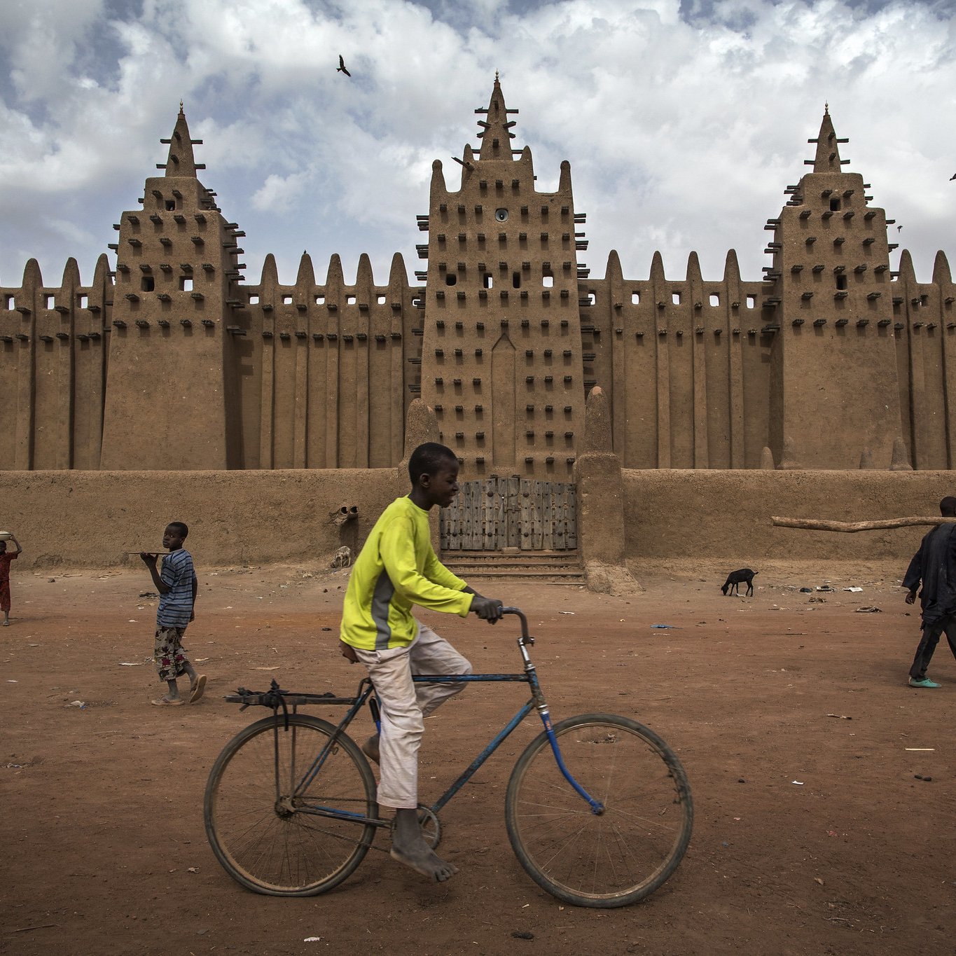 L'image montre une grande mosquée en banco, typique de l'architecture soudanaise, avec des murs en terre crue qui s'élèvent vers le ciel. Sa façade est ornée de nombreuses pointes et créneaux, formant un motif impressionnant. Au premier plan, un jeune garçon traverse la scène à bicyclette, tandis que d'autres enfants jouent et se déplacent dans l'espace. Le sol est poussiéreux, et le climat semble chaud, avec des nuages ici et là dans le ciel. L'ambiance est vivante, illustrant une scène animée de la vie quotidienne.