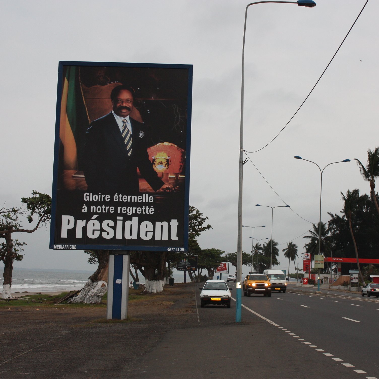 L'image montre un grand panneau publicitaire sur le bord d'une route. Ce panneau présente le portrait d'un homme en costume, avec un air solennel, tenant un insigne ou un symbole de pouvoir. En dessous de son image, il y a un texte qui dit : "Gloire éternelle à notre regretté Président", exprimant un hommage à un dirigeant décédé. En arrière-plan, on peut apercevoir une route avec quelques voitures et des palmiers, ainsi qu'une plage visible à proximité. Le ciel est nuageux, ce qui donne une ambiance mélancolique à la scène.