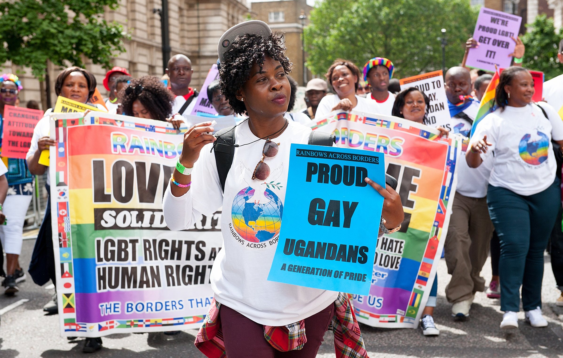 Sur cette image, un groupe de personnes marche ensemble lors d'une manifestation. Au premier plan, une femme souriante porte un t-shirt blanc avec un logo coloré représentant un globe. Elle tient un panneau bleu sur lequel il est écrit "PROUD GAY UGANDANS" en lettres grandes et claires. En arrière-plan, d'autres participants portent divers panneaux affichant des messages de solidarité et de revendication des droits LGBTQ+. La scène est animée et colorée, avec des drapeaux et des tenues qui soulignent la diversité et la fierté. L'ambiance est festive, mais également engagée, reflétant une atmosphère de lutte pour les droits humains.