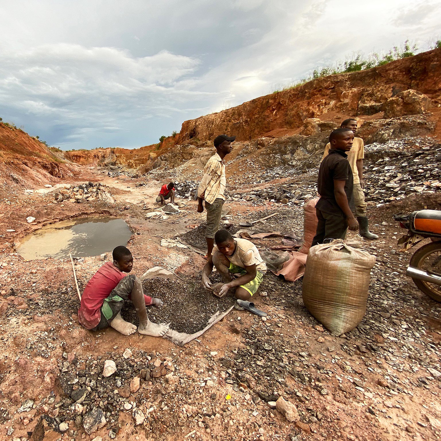 L'image montre une scène rurale d'exploitation minière. On y voit un paysage marqué par des fouilles, avec un sol rouillé et des montagnes de terre séparées. Au premier plan, plusieurs personnes, principalement des hommes et des jeunes garçons, sont en train de travailler. Certains sont accroupis, tamisant des pierres ou ramassant des minéraux, tandis que d'autres semblent transporter des sacs pleins de matériaux. À gauche, il y a un petit plan d'eau, qui est trouble, entouré de débris miniers. Le ciel est nuageux, ajoutant une ambiance sombre à la scène, évoquant un travail difficile et épuisant.