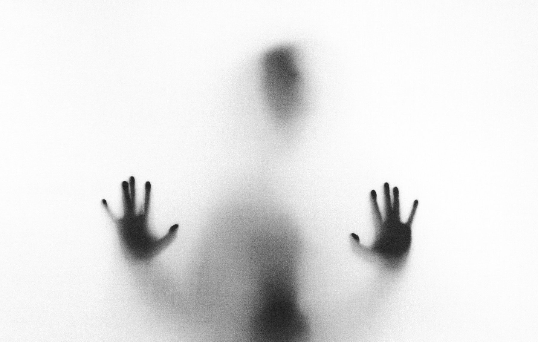 L'image représente une silhouette floue d'une personne, en arrière-plan léger. La figure se trouve proche d'une surface translucide, créant un effet de brouillard. On peut distinguer des mains, les paumes dirigées vers l'avant, comme si la personne essayait de pousser ou de traverser cette barrière. Les contours sont indistincts, évoquant une sensation de mystère et d'isolement. L'ambiance générale est à la fois intrigante et un peu mélancolique.