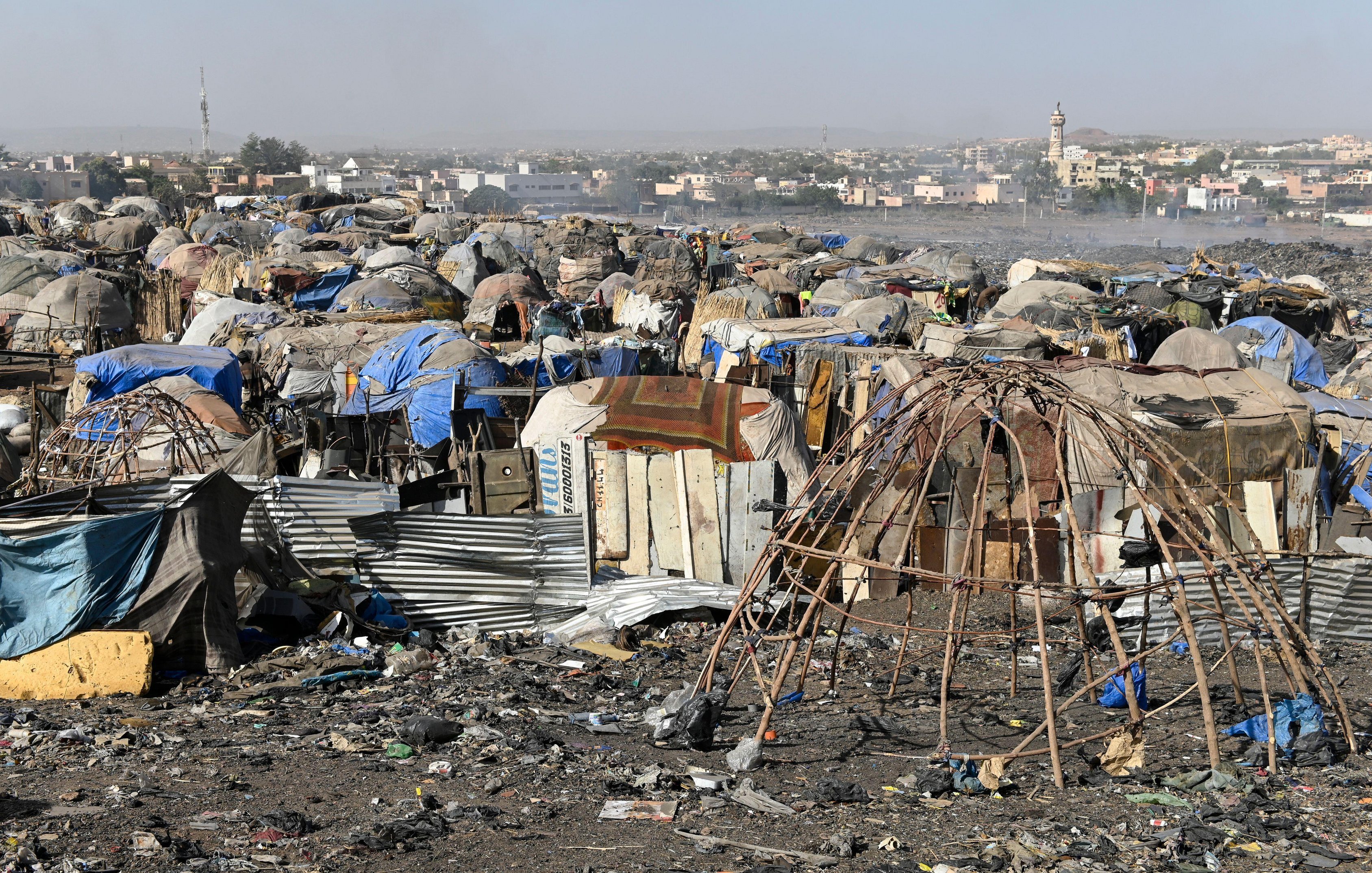 L'image montre un paysage de bidonville, où l'on peut distinguer des habitations précaires construites à partir de matériaux recyclés tels que des tôles, des bâches en plastique et des branches de bois. Ces structures sont souvent assez rudimentaires et forment des petites communautés. Le sol est jonché de déchets et de débris divers, reflétant une forte pollution. À l'arrière-plan, on aperçoit des bâtiments en dur, ainsi qu'une mosquée avec son minaret, indiquant la présence d'une ville proche. L'atmosphère dégage une impression de désolation, mais aussi de vie, avec des traces humaines visibles malgré les conditions difficiles.