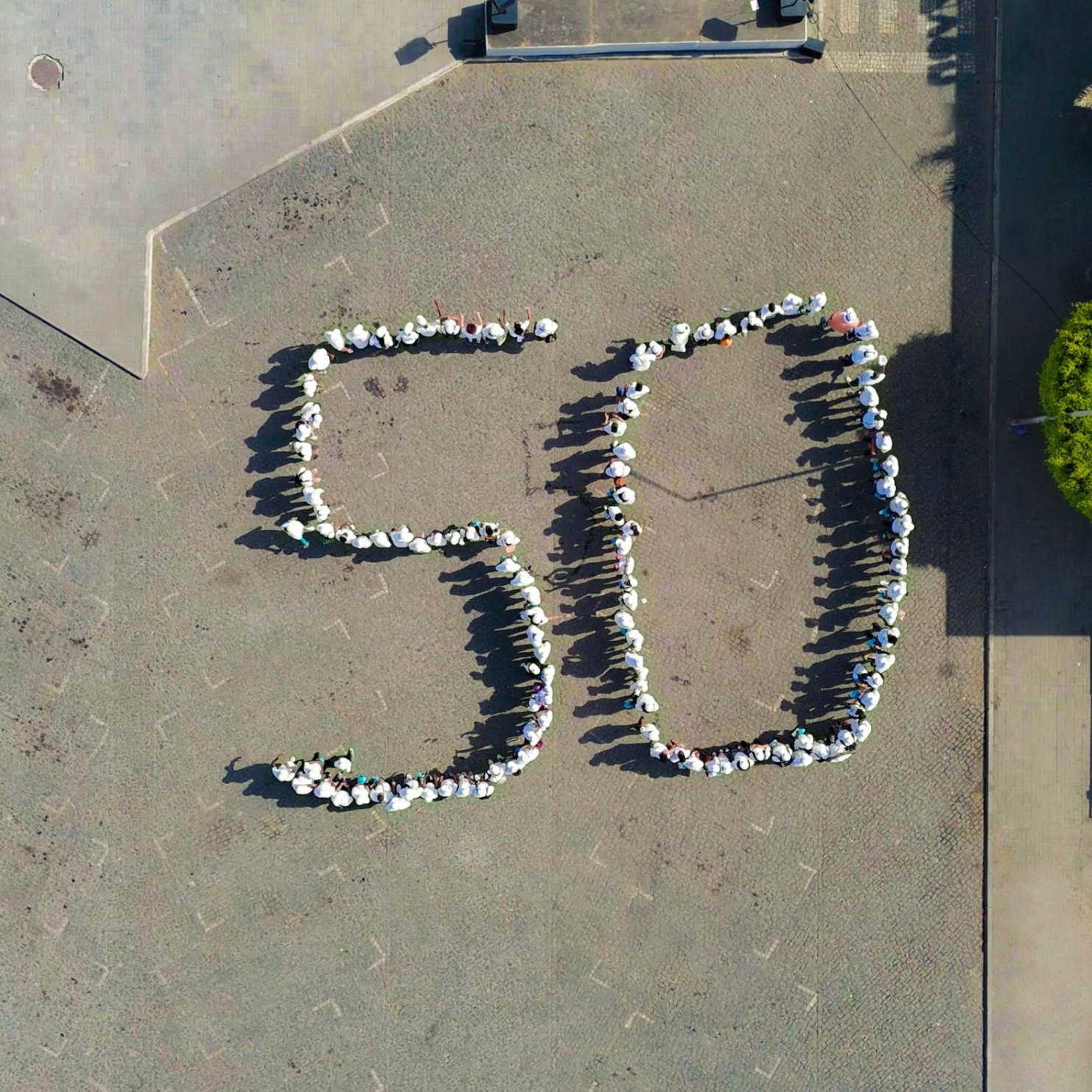 L'image montre une vue aérienne d'une place où des personnes sont réunies pour former le chiffre "50". Les participants sont disposés de manière à créer les deux chiffres avec leurs corps, et ils semblent porter des vêtements clairs, peut-être des t-shirts blancs. Autour, on peut voir des pavés au sol et quelques éléments de paysage urbain, comme des arbres et des bâtiments en arrière-plan. L'ambiance semble conviviale et festive, suggérant que cet événement marque une célébration ou un anniversaire.