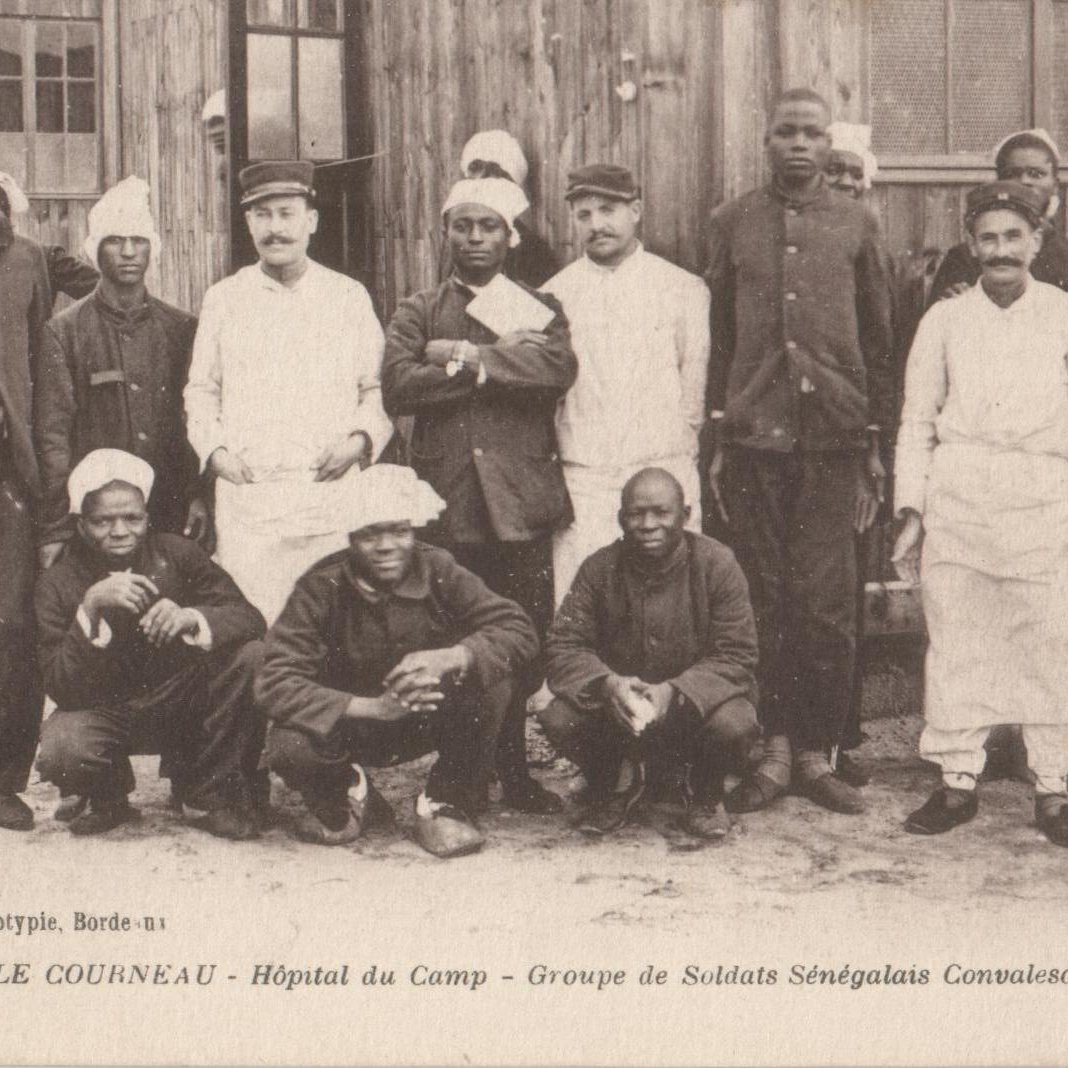 L'image montre un groupe de soldats sénégalais convalescents rassemblés devant un bâtiment en bois, vraisemblablement un hôpital de campagne. Il y a treize personnes, dont des hommes en uniforme militaire et des membres du personnel médical. Les soldats portent des uniformes sombres et certains d'entre eux ont des casquettes ou des bonnets. Le personnel médical, d'autre part, porte des blouses blanches, indiquant leur rôle. Tous affichent des expressions amicales, certains sourient tandis que d'autres prennent des poses plus sérieuses. L'arrière-plan montre des murs en bois, donnant une atmosphère de campagne ou de mobilisation militaire. Cette scène capture un moment de camaraderie et de résilience au sein des soldats pendant une période difficile.