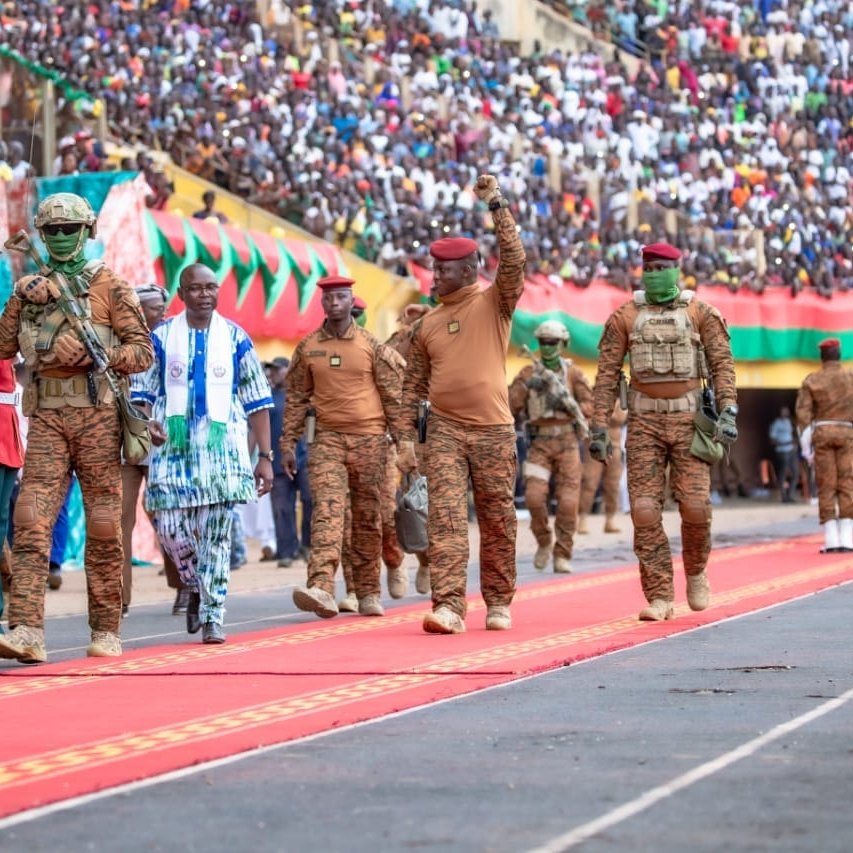 L'image dépeint une scène d'événement public dans un stade. Un grand nombre de personnes est rassemblé dans les gradins, créant une ambiance festive et patriotique. Au premier plan, des militaires en uniforme brun avancent sur un tapis rouge, symbolisant une cérémonie officielle. L'un des militaires, portant une casquette rouge, lève le bras en signe de salutation. À ses côtés, un homme en vêtements traditionnels aux motifs colorés marche avec assurance. La scène est remplie de drapeaux et de banderoles qui ajoutent des touches de couleur à l'événement. L'atmosphère est vibrante, marquée par l'engagement et l'unité.