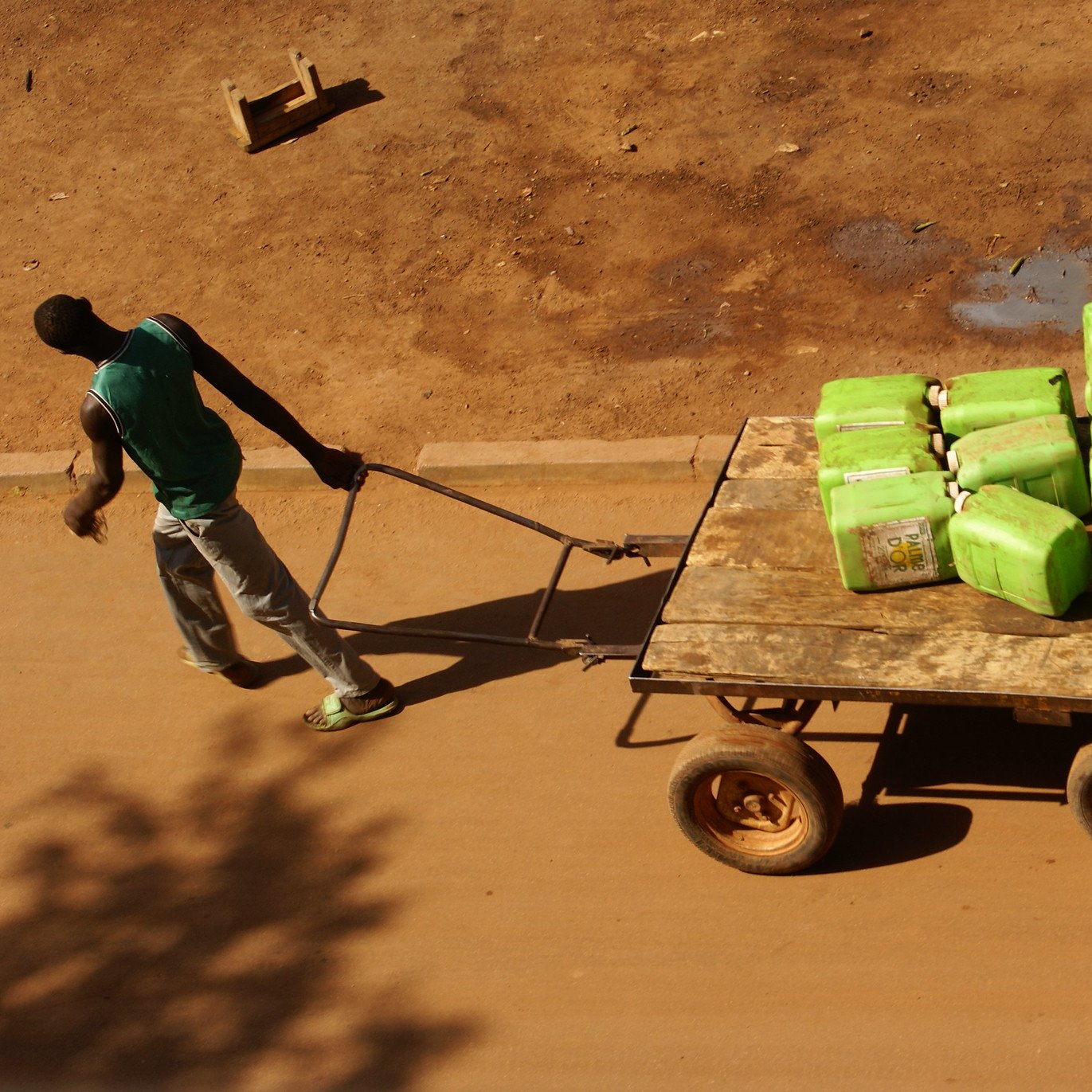 L'image montre un homme poussant une charrette en bois sur une route en terre. La charrette est remplie de nombreuses bouteilles vertes, probablement des contenants d'eau. L'homme porte un T-shirt sans manches et un pantalon gris, et son corps semble penché en avant, indiquant l'effort physique qu'il fournit. Le sol autour de lui est sec et légèrement poussiéreux, avec quelques traces visibles montrant que d'autres véhicules ou personnes ont déjà passé. En arrière-plan, il pourrait y avoir des objets comme des seaux ou des tuyaux, mais l'accent est mis sur l'homme et sa cargaison. La lumière est forte, suggérant une journée ensoleillée.
