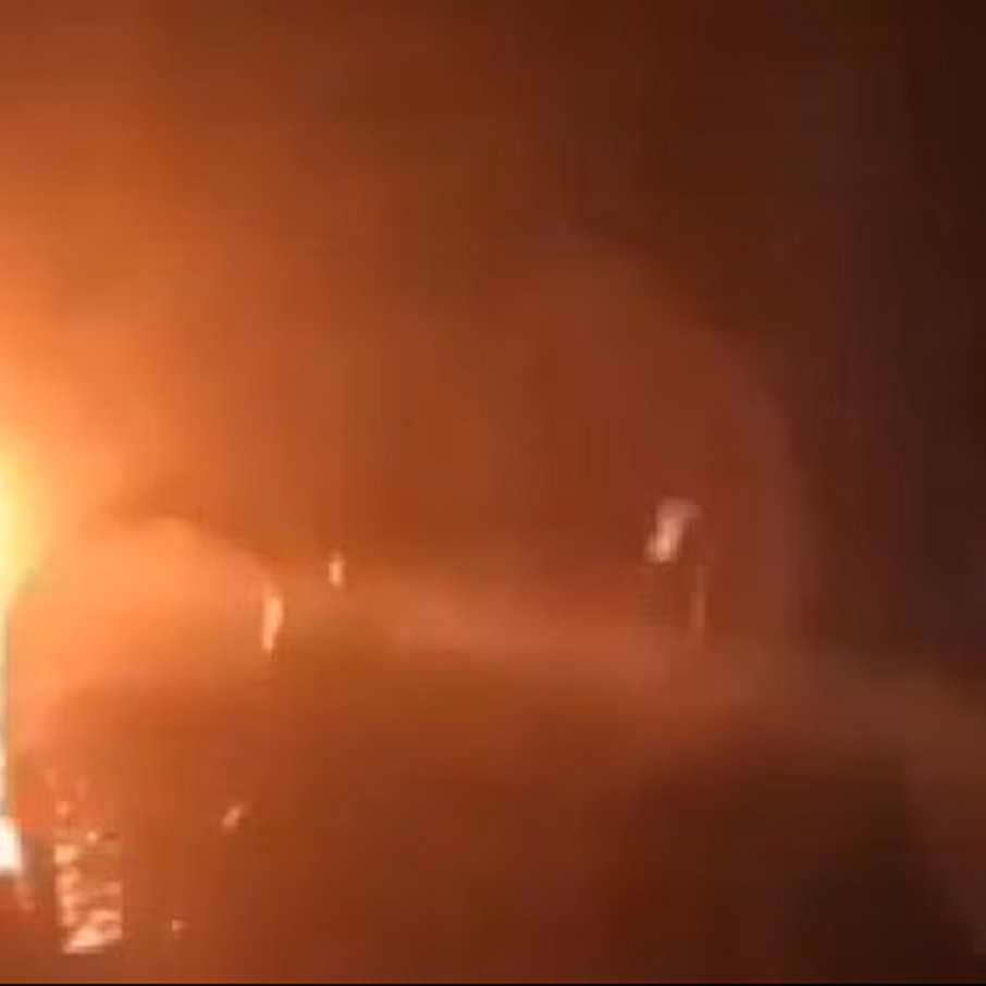 The image appears to show a bright light source in a dark environment, suggesting a fire or an explosion. The intense glow creates a dramatic contrast against the dark background, and there are vague silhouettes or shapes surrounding the light, which could indicate the presence of objects or people nearby. The overall atmosphere seems tense and chaotic due to the brightness and obscurity of the other elements in the scene.