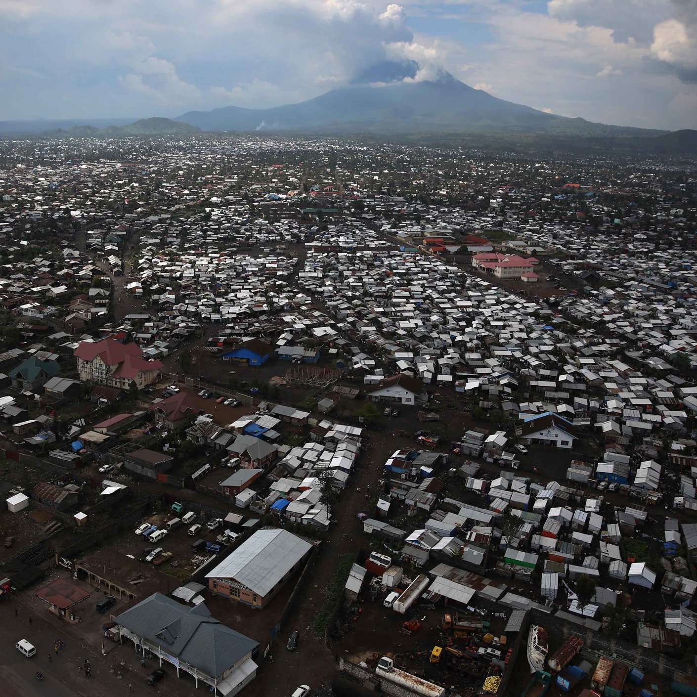 Imaginez une vaste étendue urbaine vue d'en haut, où des milliers de toits en tôle ondulée et en matériaux divers se mêlent pour créer un paysage dense. Les bâtiments semblent serrés les uns contre les autres, formant un réseau labyrinthique. Au loin, un majestueux volcan émerge à l'horizon, avec des sommets nuageux qui soulignent sa présence imposante. Le ciel présente des teintes variées de gris et de bleu, projetant une lumière diffuse sur la scène. En bas, des routes sont visibles, animées de véhicules et de personnes, ajoutant une sensation de vie à cet environnement. Cette image évoque à la fois la densité urbaine et la force tranquille de la nature en toile de fond.