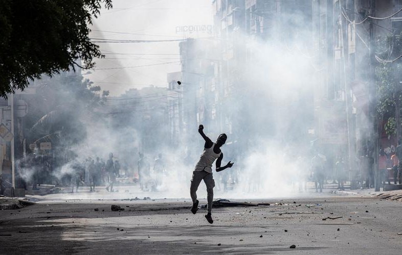 L'image dépeint une scène urbaine troublée, où la tension est palpable. Au premier plan, une silhouette humaine, probablement un homme, est en mouvement, lançant un projectile. Il semble néanmoins être entouré de foules, suggérant une situation tumultueuse. Des nuages de fumée épaisse s'élèvent dans l'air, créant une atmosphère de chaos, tandis que des débris jonchent le sol, témoignant d'une agitation récente. En arrière-plan, on peut deviner des bâtiments urbains flous, et la lumière est sombre, renforçant l'impression d'une émeute ou d'une protestation. Les sons et odeurs d'une ville agitée pourraient également accompagner cette image, avec des cris, des sirènes, ou l'odeur de fumée.