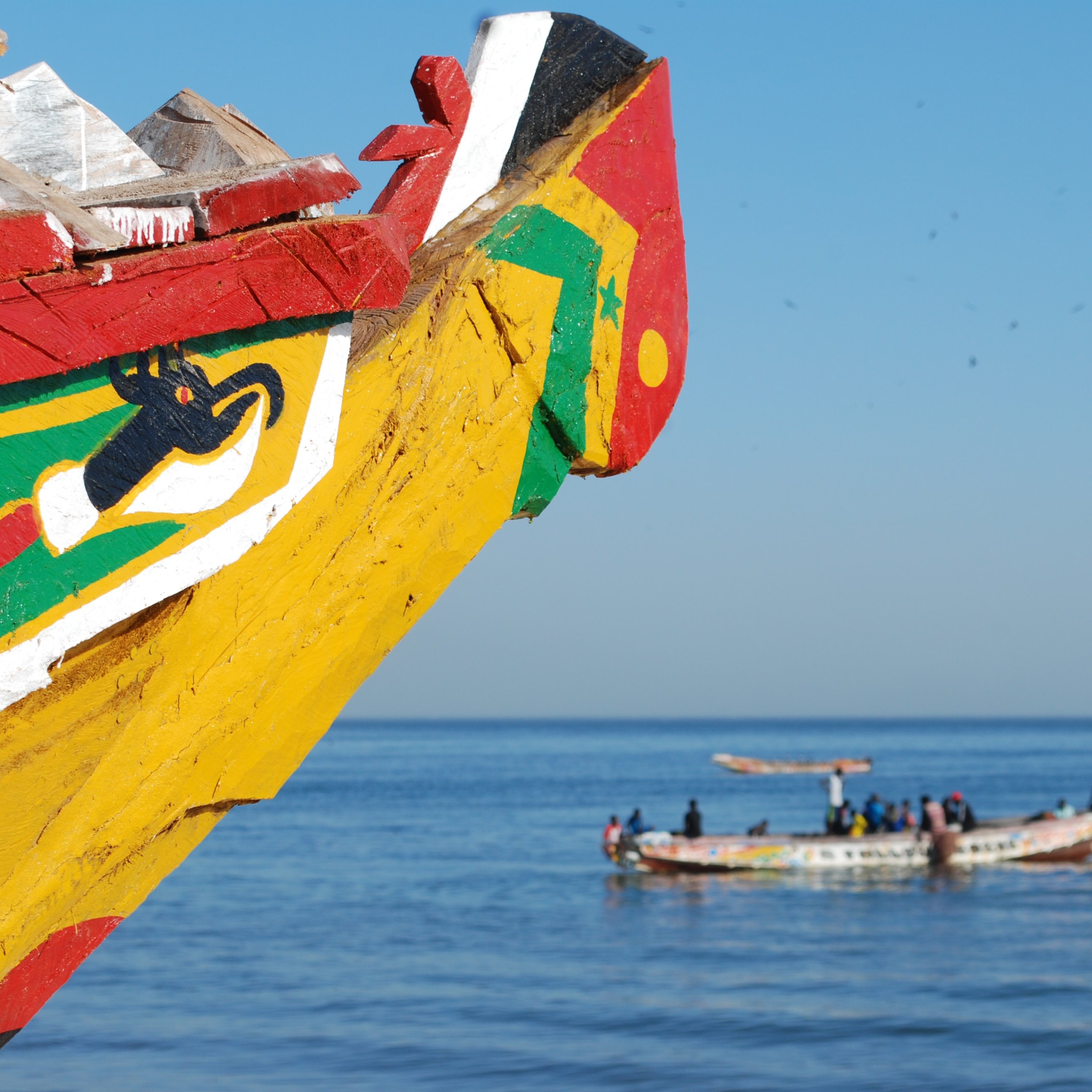 Cette image montre la proue d'une embarcation colorée, ornée de motifs vive avec principalement des couleurs rouge, jaune et verte. Les dessins évoquent des éléments symboliques, peut-être liés à la culture locale. En arrière-plan, on aperçoit d'autres bateaux sur l'eau, avec des personnes à bord, travaillant ou naviguant paisiblement. La mer apparaît calme, reflétant un ciel bleu sans nuages, créant une atmosphère sereine et ensoleillée.
