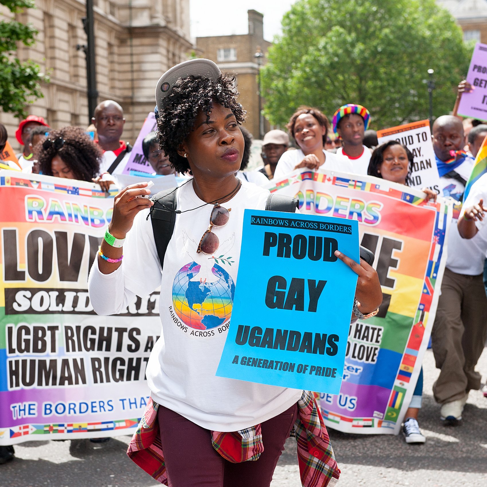 Sur cette image, un groupe de personnes marche ensemble lors d'une manifestation. Au premier plan, une femme souriante porte un t-shirt blanc avec un logo coloré représentant un globe. Elle tient un panneau bleu sur lequel il est écrit "PROUD GAY UGANDANS" en lettres grandes et claires. En arrière-plan, d'autres participants portent divers panneaux affichant des messages de solidarité et de revendication des droits LGBTQ+. La scène est animée et colorée, avec des drapeaux et des tenues qui soulignent la diversité et la fierté. L'ambiance est festive, mais également engagée, reflétant une atmosphère de lutte pour les droits humains.