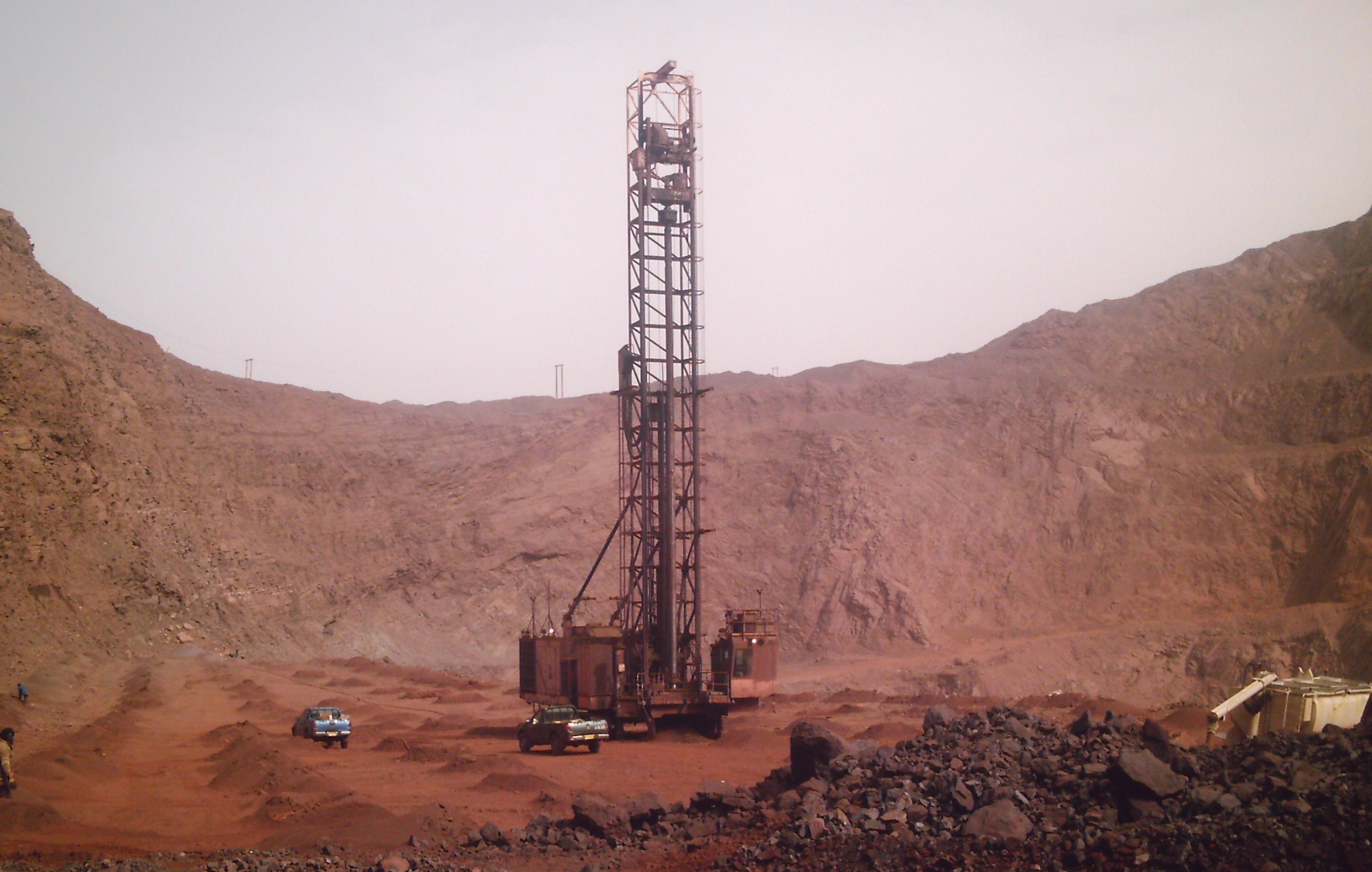 L'image montre un site d'exploitation minière dans un environnement aride et rocheux. Au centre, une grande foreuse se dresse, entourée de montagnes de terre et de roches. La foreuse, de couleur rouille, a une structure métallique imposante qui semble clé dans le processus d'extraction. Autour d'elle, plusieurs véhicules, probablement des camions ou des 4x4, sont garés sur le sol. Le terrain environnant est rocailleux et dépouillé, avec des teintes ocres et brunes, évoquant une atmosphère sévère et industrielle. Le ciel est clair, ce qui accentue l'impression de chaleur et d'aridité de cette zone minière.