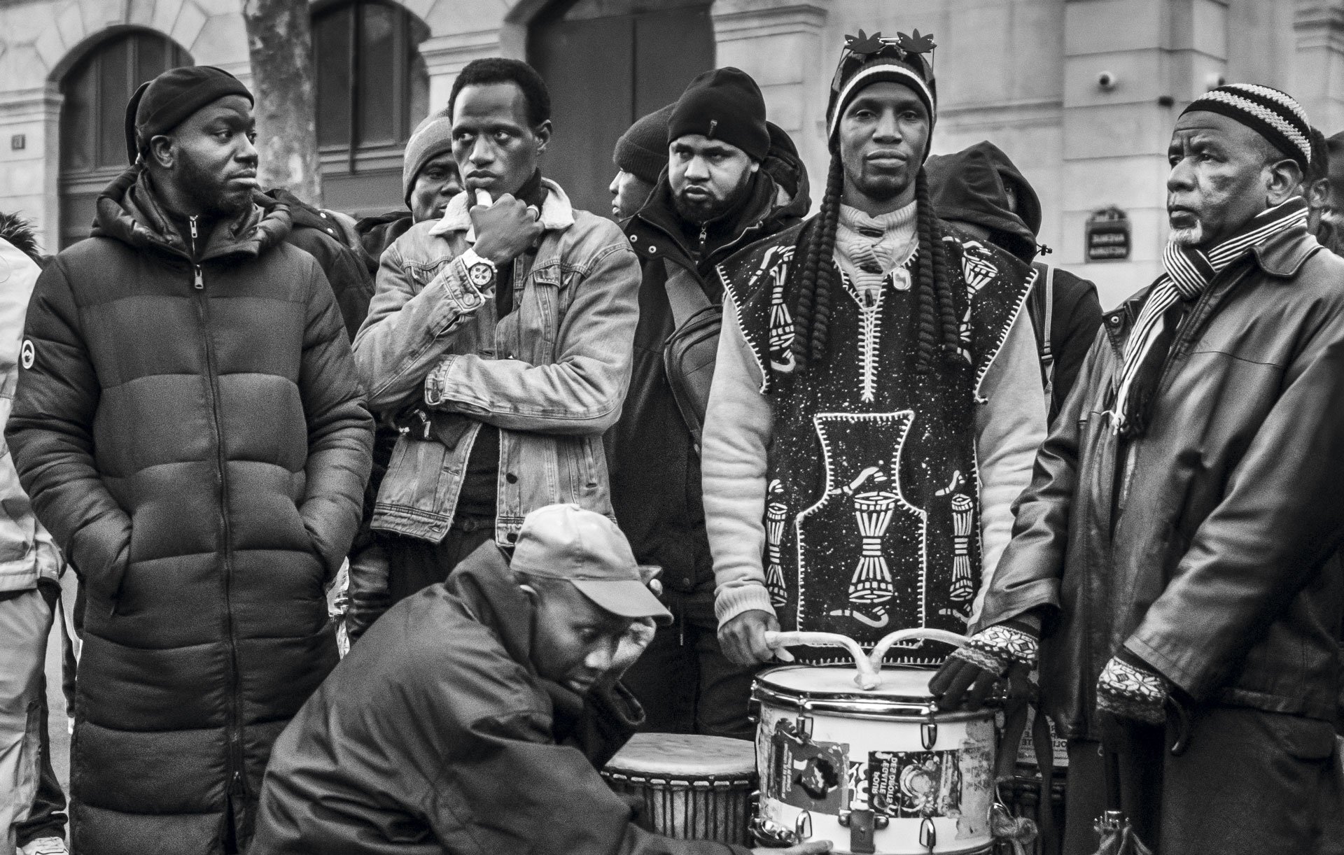 L'image montre un groupe de plusieurs hommes rassemblés dans un environnement urbain. Ils portent des vêtements variés, allant de parkas sombres à des vêtements traditionnels. Certains d'entre eux regardent dans des directions différentes, affichant une diversité d'expressions. Au centre, un homme est assis, jouant sur un tambour, ajoutant une ambiance musicale à la scène. Les autres semblent attentifs, créant une atmosphère de camaraderie et d'échange. L'image est en noir et blanc, accentuant les contrastes et les textures des vêtements et des visages.