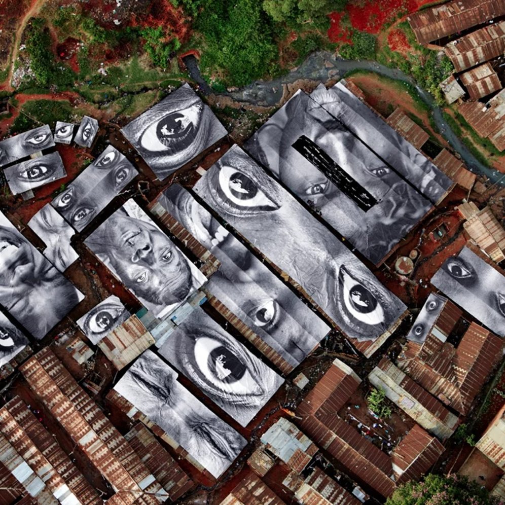 L'image représente une vue aérienne d'un bidonville, où les toits en tôle ondulée sont de différentes teintes, principalement marron. Sur ces toits, il y a de grandes œuvres d'art en noir et blanc illustrant des visages humains. Les yeux des portraits semblent fixer le spectateur, créant une impression d'intensité et de connexion. Entre les toits, on aperçoit des espaces plus clairs, peut-être des chemins ou des ruelles, et on peut deviner la verdure environnante qui ajoute une touche de contraste à l'environnement urbain. L'ensemble évoque à la fois la pauvreté du lieu et la richesse des émotions humaines capturées dans les portraits.