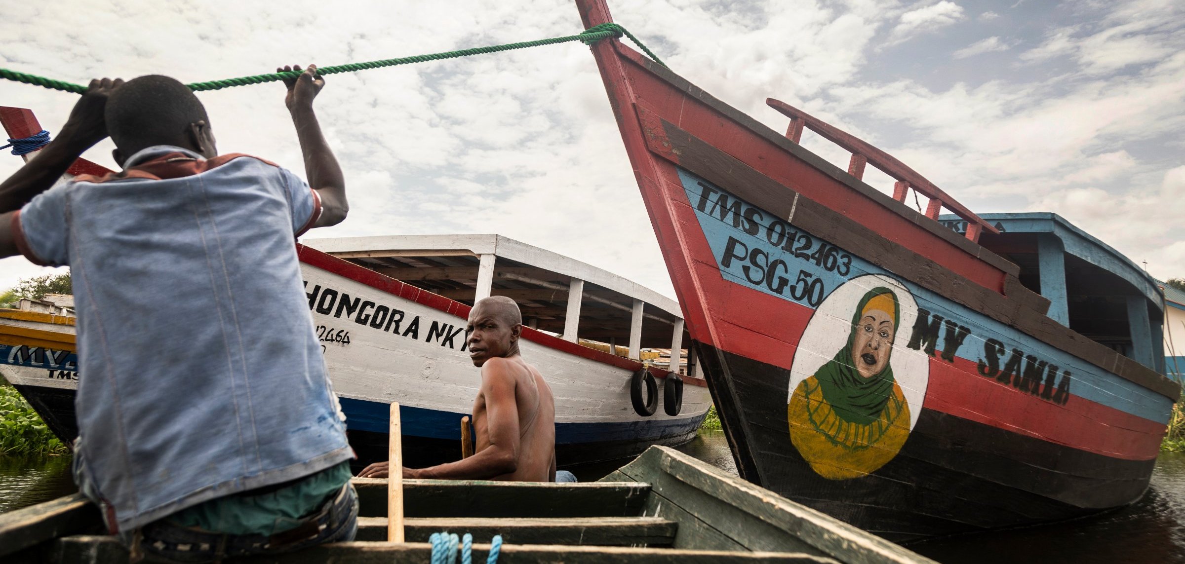L'image montre deux bateaux colorés amarrés au bord d'un cours d'eau calme. À l'avant, un homme, en train de ramer, est visible, concentré sur son effort. Un autre homme est assis à l'arrière, regardant vers l'objectif. Les bateaux sont peints de manière vive, avec des inscriptions comme "MY SAID" et "MONGORA MK". L'un des bateaux présente également une grande peinture d'un visage souriant. Le ciel est partiellement nuageux, ajoutant une dimension lumineuse à la scène. L'atmosphère est paisible, évoquant une ambiance de vie flottante.