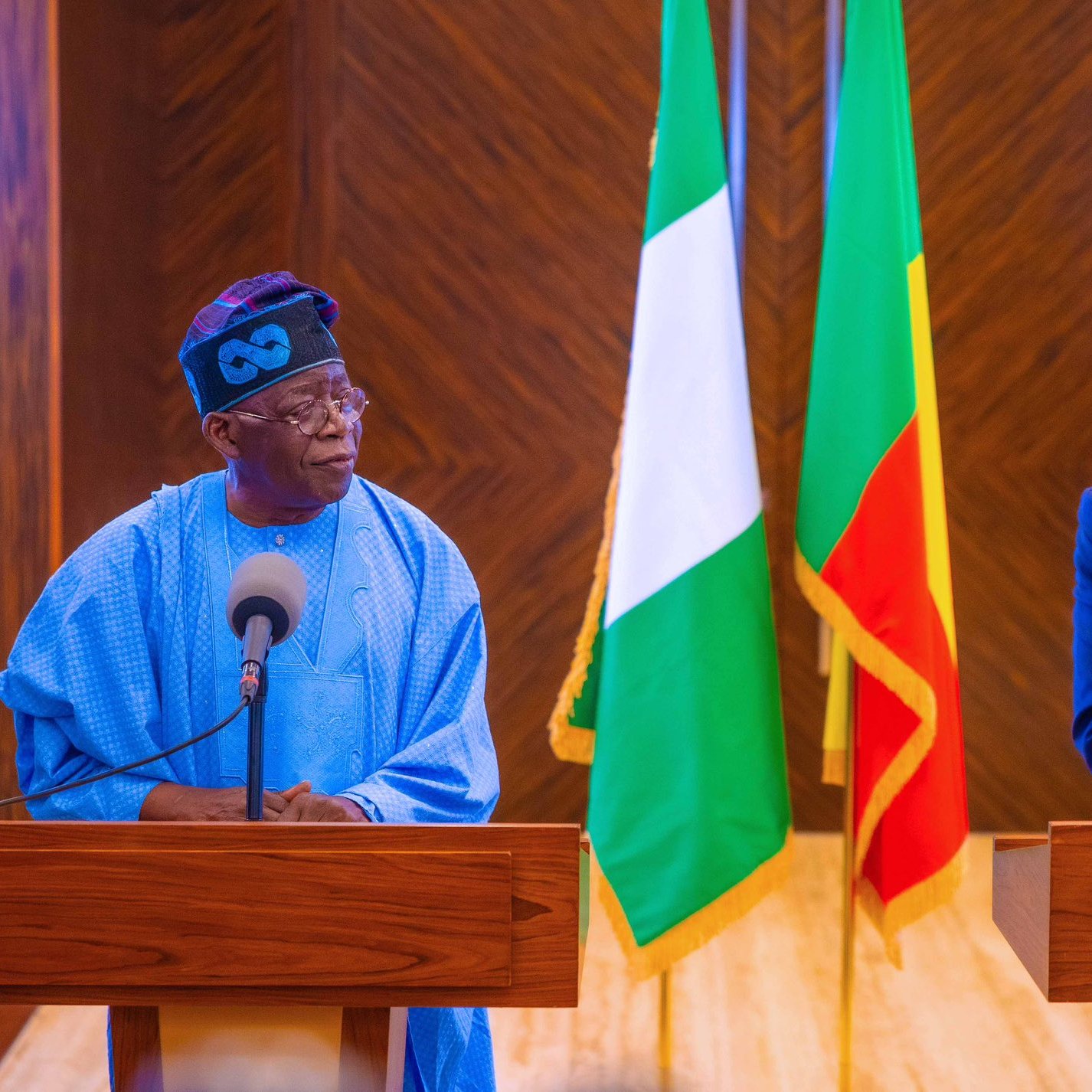 L'image montre deux hommes se tenant devant des pupitres en bois dans un cadre officiel. À leur arrière-plan, on peut voir les drapeaux du Nigéria (vert, blanc, vert) et du Bénin (rouge, jaune, vert). L'homme à gauche porte une tenue traditionnelle de couleur bleue avec un chapeau, tandis que l'homme à droite est habillé en costume sombre. Les deux hommes semblent engagés dans une conversation, exprimant une atmosphère de dialogue et de coopération. Le décor est élégant, soulignant le caractère officiel de la rencontre.