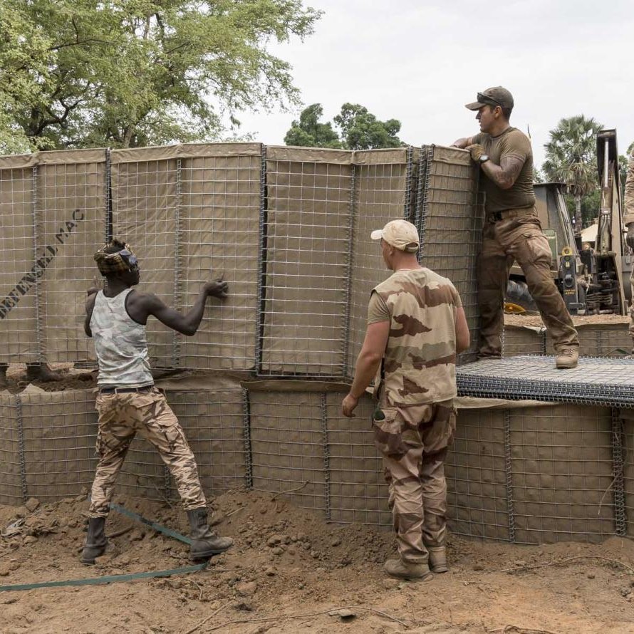 L'image montre un groupe de soldats au travail sur un site de construction. Ils sont en train de manipuler de grandes structures en treillis, qui semblent être remplies de terre. L'environnement est naturel, avec des arbres verts en arrière-plan et un sol légèrement argileux. Les soldats portent des uniformes militaires et semblent concentrés sur leur tâche. Certains d'entre eux soulèvent des éléments, tandis que d'autres les positionnent soigneusement. Cette scène évoque une atmosphère de collaboration et d'effort collectif.