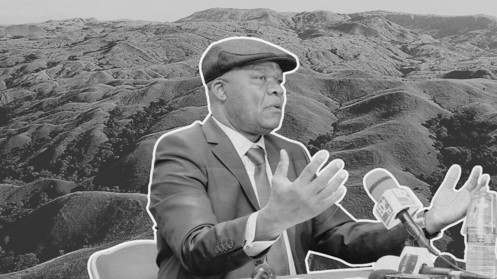 L'image montre un homme en costume, avec un chapeau, en train de parler lors d'une conférence ou d'une interview. Il est assis devant un microphone et semble gesticuler pour appuyer ses propos. En arrière-plan, on aperçoit un paysage de collines ondulantes, évoquant une nature sauvage et montagneuse, donnant une impression de profondeur et de calme. L'ensemble de l'image est en noir et blanc, ce qui renforce le sérieux de la scène.