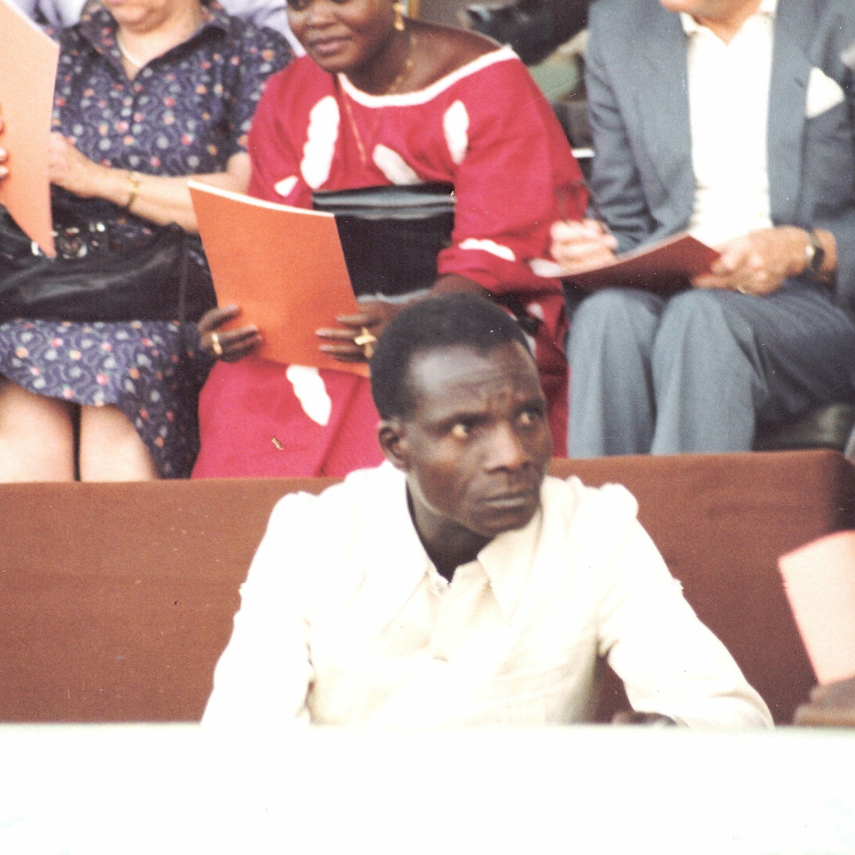 L'image montre une scène d'un événement où un homme en chemise claire est assis au premier plan, semblant sérieux et attentif. À l'arrière-plan, plusieurs personnes discutent, certaines tenant des documents ou des livrets. On aperçoit des vêtements colorés, peut-être d'origine africaine, ce qui ajoute une dimension culturelle à la scène. L'atmosphère générale évoque un rassemblement formel ou une cérémonie.