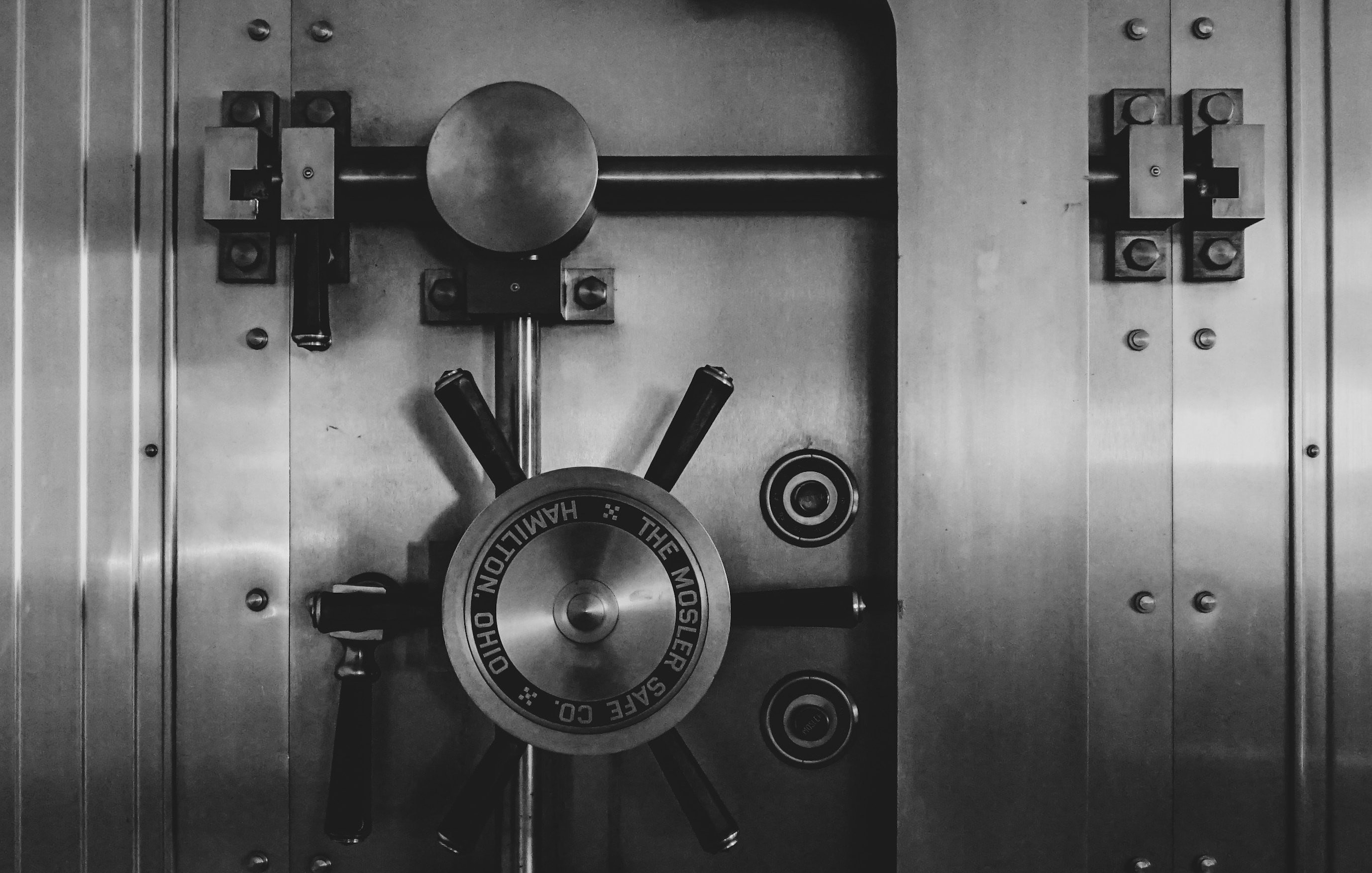 Imaginez une grosse porte de coffre-fort, conçue pour être très sécurisée. La surface est en métal brillant, avec des nuances de gris et une texture lisse au toucher. Au centre, il y a un gros volant, un peu comme un cercle en métal, qui permet de verrouiller ou déverrouiller la porte. Ce volant présente des inscriptions le long de son diamètre, indiquant probablement le nom du fabricant. De chaque côté, il y a des verrous et des mécanismes qui ajoutent à l'impression de robustesse et de sécurité. L'ensemble dégage une sensation de solidité et de mystère, comme si de précieux trésors étaient cachés derrière cette porte imposante.