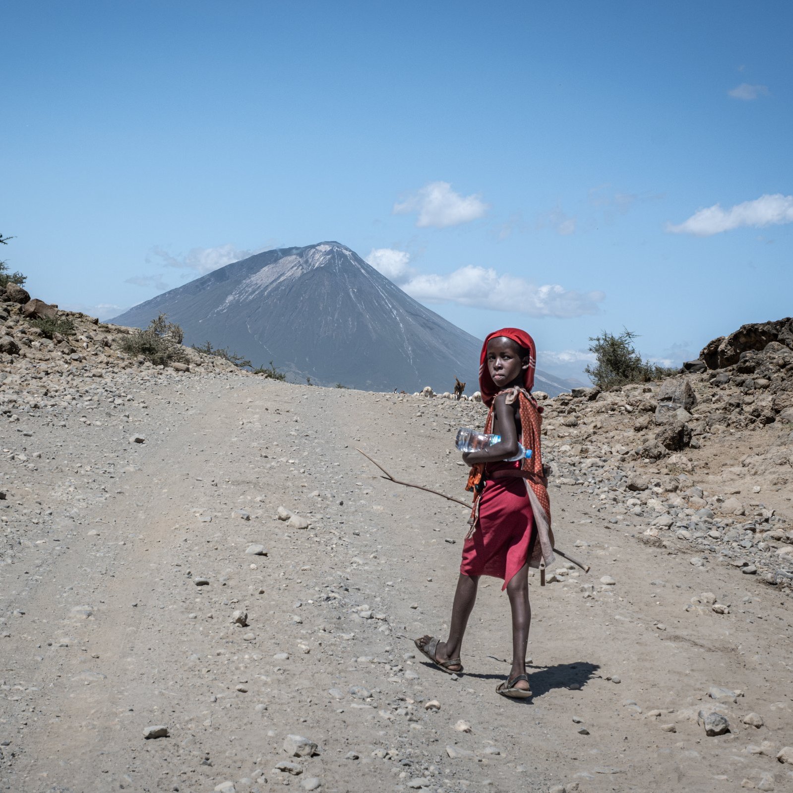 L'image montre un enfant marchant sur un chemin de terre. Il porte une robe rouge et un foulard, et il semble porter un petit récipient ou une bouteille dans ses mains. En arrière-plan, on aperçoit un volcan majestueux, dont le sommet est légèrement nuageux sous un ciel bleu clair parsemé de quelques nuages blancs. Le terrain autour est rocailleux et aride, avec quelques petits buissons éparpillés. L'ensemble de la scène dégage une atmosphère de nature sauvage et éloignée.