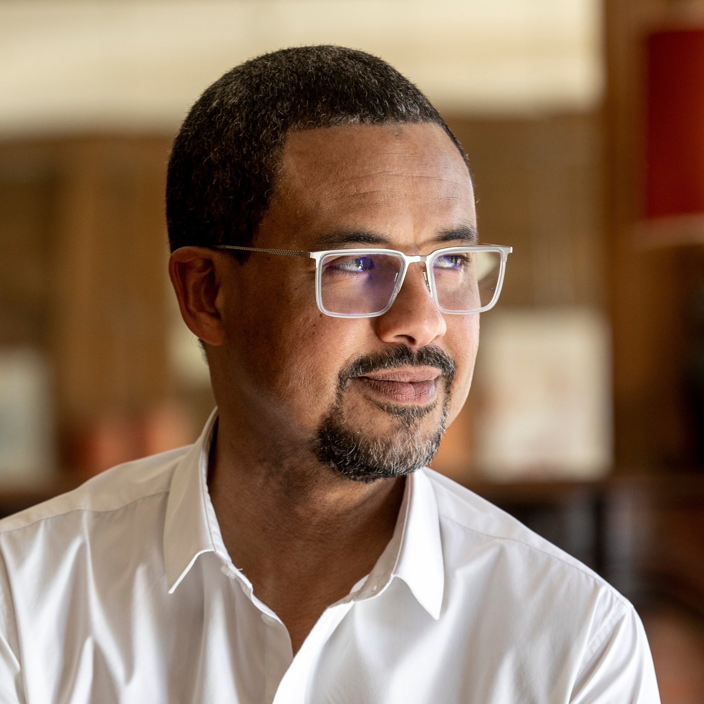 L'image présente un homme d'âge moyen, debout légèrement sur le côté. Il a la peau foncée et porte des lunettes avec une monture claire. Son expression est contemplative, avec un léger sourire aux lèvres. Il porte une chemise blanche, qui contraste avec son teint. En arrière-plan, on peut apercevoir des lampes avec des abat-jour orange et un décor en bois qui donne une ambiance chaleureuse et intime à l'ensemble. La lumière est douce, créant une atmosphère paisible.