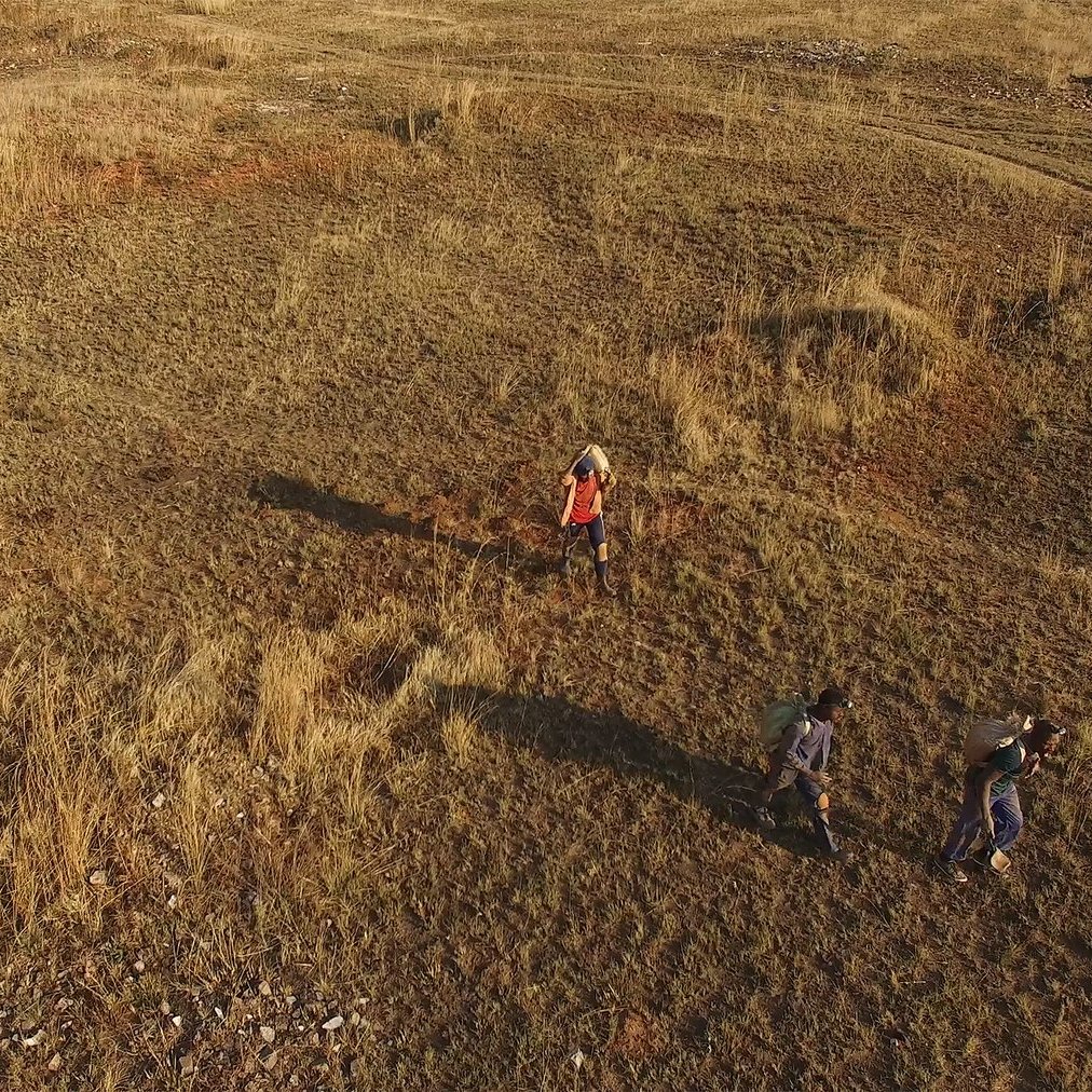 Dans cette image, on voit un paysage vaste et ouvert, couvert d'herbe sèche et de petites touffes de plantes. Le sol est légèrement accidenté, avec des zones de terre claire et quelques pierres éparpillées. Trois personnes marchent dans ce terrain. L'une d'elles porte un vêtement rouge, tandis que les autres sont habillées de couleurs plus sombres. Leur ombre s'étend sur le sol, suggérant que le soleil est assez bas dans le ciel. L'atmosphère est calme et naturelle, évoquant une promenade en pleine nature.