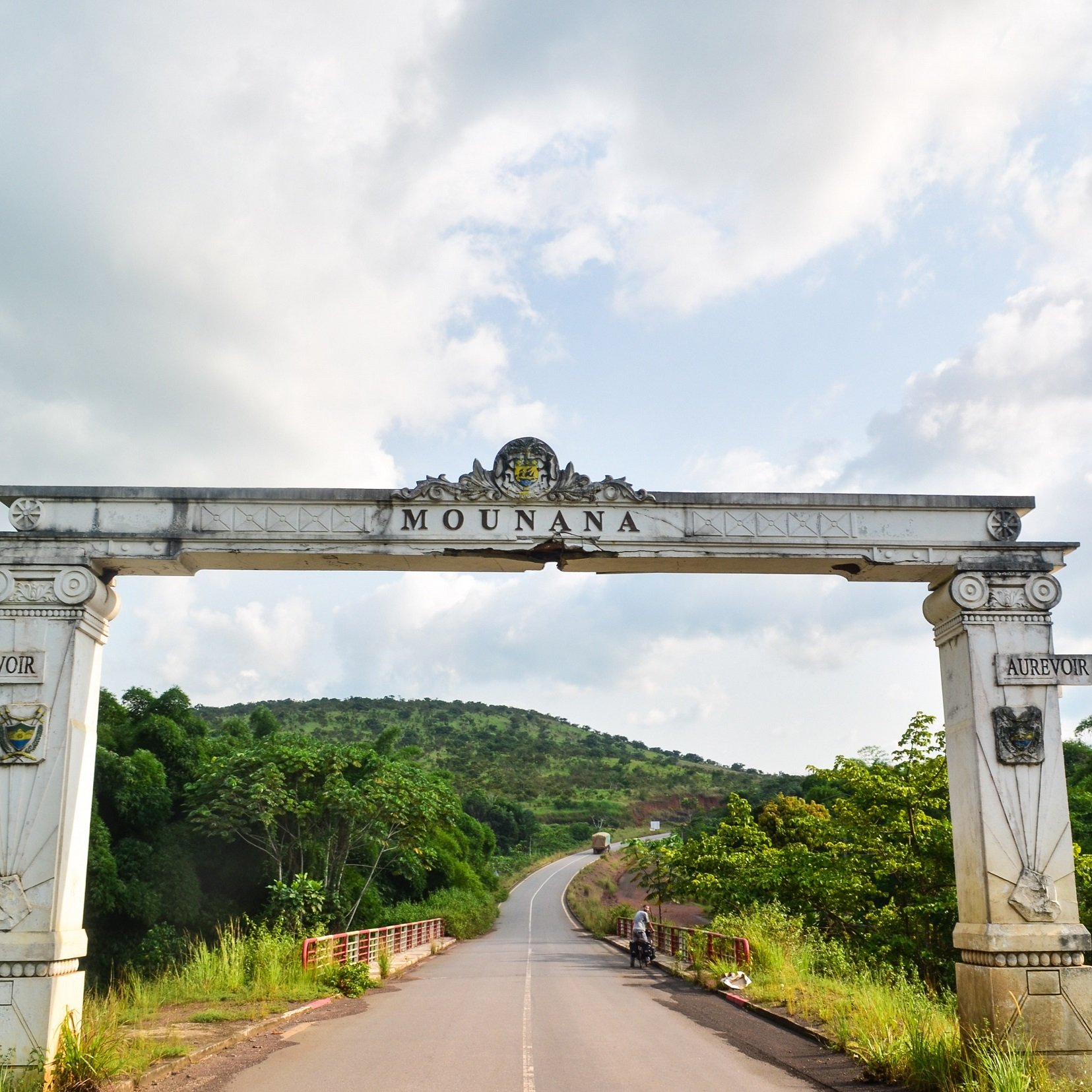 L'image présente une grande arche située à l'entrée de la ville de Mounana. Cette arche, ornée de motifs architecturaux, est peinte en blanc avec des détails dorés et armoriaux de chaque côté. Au-dessus de l'entrée, le nom "MOUNANA" est inscrit en lettres stylisées. En arrière-plan, on aperçoit un paysage verdoyant avec des collines et une route qui serpente entre des arbres. Le ciel est partiellement nuageux, créant une atmosphère paisible et accueillante. L'ensemble évoque l'entrée d'un lieu significatif, invitant les visiteurs à découvrir ce qui se trouve au-delà de l'arche.