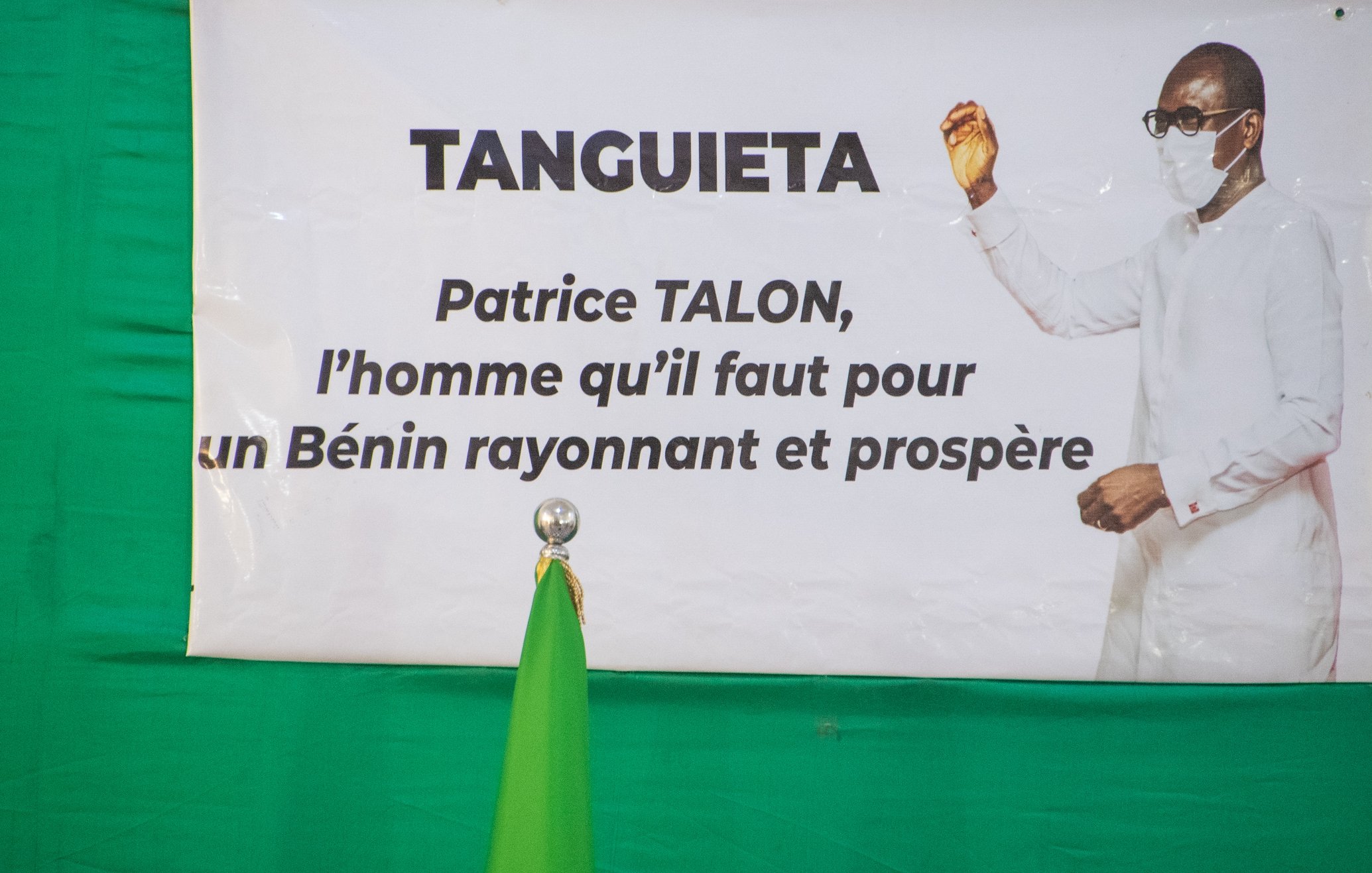 L'image présente une affiche accrochée sur un fond vert. Cette affiche met en avant le nom "TANGUIETA" en haut, suivi d'une mention concernant Patrice TALON. Le texte indique qu'il est "l'homme qu'il faut pour un Bénin rayonnant et prospère." On peut également voir un homme en tenue blanche, portant un masque. Il est positionné à droite de l'affiche, levant une main dans un geste de discours ou de présentation. À gauche de l'affiche, se trouve un drapeau vert qui se dresse à la verticale. L'ensemble véhicule un message d'espoir et de prospérité pour le pays.