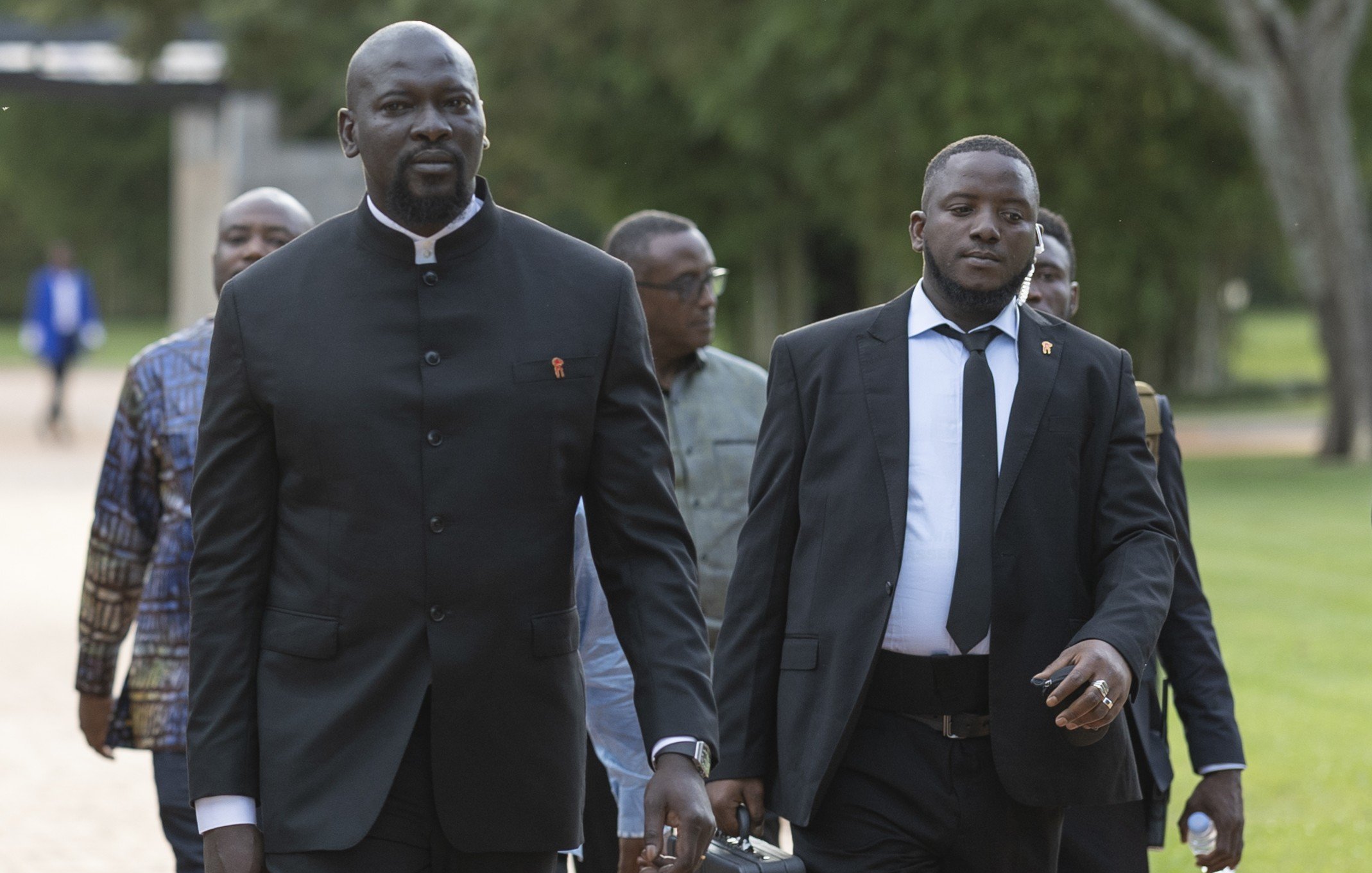L'image montre un groupe d'hommes marchant ensemble sur un chemin dans un environnement verdoyant. Au premier plan, un homme grand et imposant porte un costume noir avec un col Mao, tandis qu'un autre homme à côté de lui est en costume noir et cravate. Ils ont l'air sérieux et concentrés, comme s'ils se rendaient à un événement important. En arrière-plan, d'autres hommes sont visibles, avec des tenues variées, ce qui suggère une atmosphère de réunion ou de conférence. Le paysage est paisible, avec des arbres et une lumière douce, ajoutant à l'ambiance formelle de la scène.