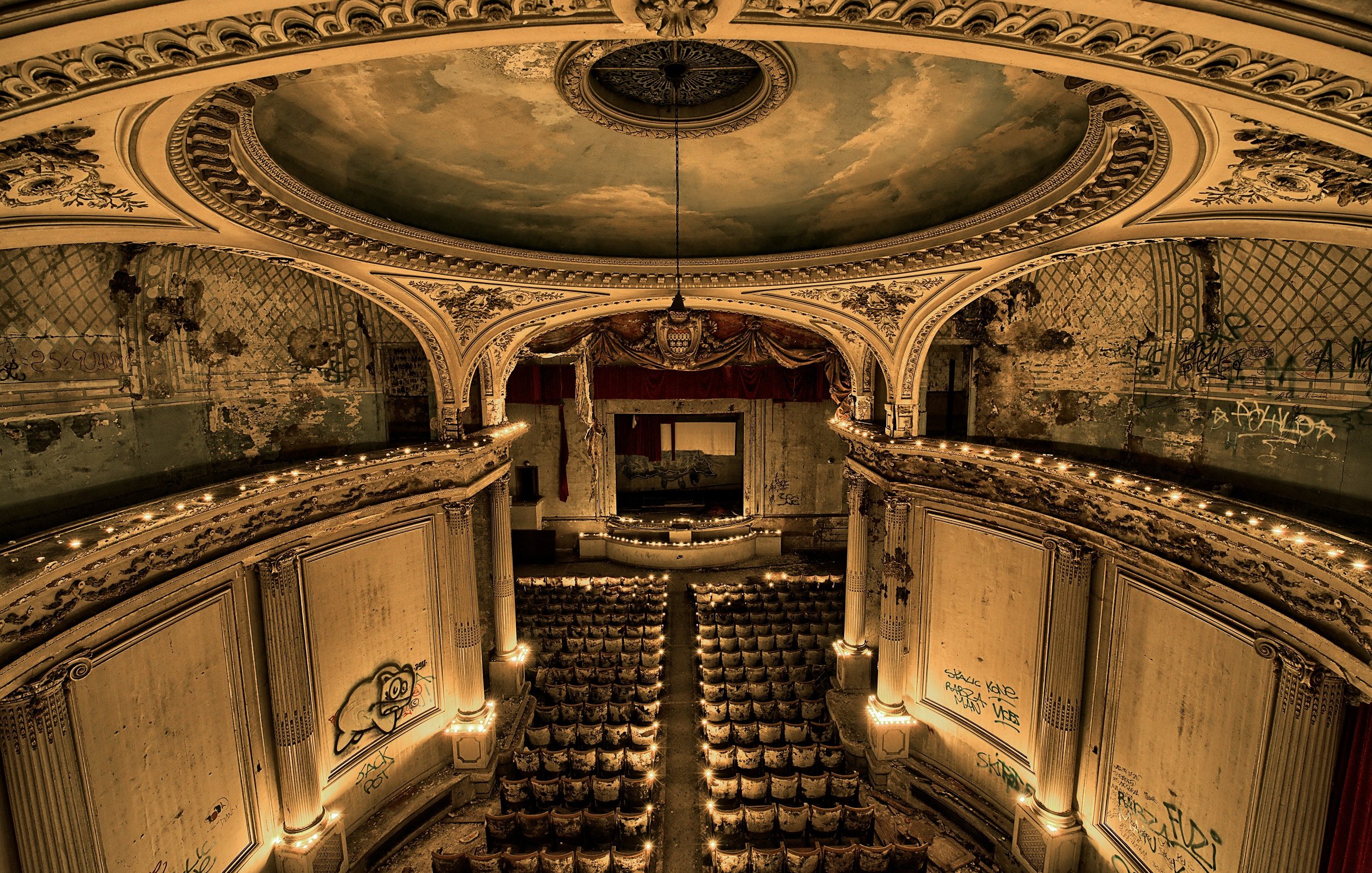 L'image représente l'intérieur d'un ancien théâtre, avec une architecture classique. Le plafond est orné de fresques délicates, tandis que les murs affichent des signes d'usure et de dégradation, témoignant du temps passé. Au centre, un grand miroir est encadré par un drapé rouge, ajoutant une touche de luxe. Devant lui, des rangées de sièges, maintenant poussiéreux, indiquent l'endroit où se tenaient les spectateurs. Des lumières tamisées créent une ambiance mélancolique, et des graffitis sur les murs rappellent l'abandon de ce lieu autrefois vivant. L'ensemble dégage une atmosphère nostalgique, marquée par la beauté d'antan et le passage du temps.