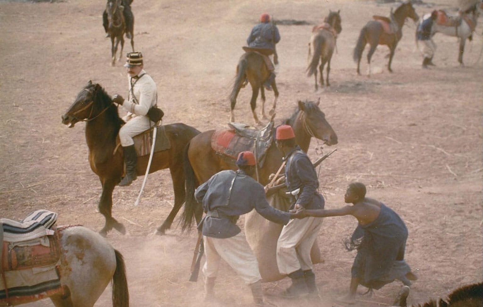 L'image présente une scène historique en plein air, où plusieurs personnages sont engagés dans une interaction dynamique. Au centre, un homme vêtu d'un uniforme militaire blanc est assis sur un cheval, observant la scène. Deux autres hommes, probablement des soldats, sont en train de se battre ou de grappler au sol, mettant en évidence un moment de tension. En arrière-plan, plusieurs autres chevaux et hommes se déplacent dans un environnement poussiéreux, suggérant une ambiance mouvementée et chaotique, typique d'un camp militaire. Le terrain est sec, peut-être de la terre battue, ce qui ajoute à l'atmosphère rustique et historique de la scène.