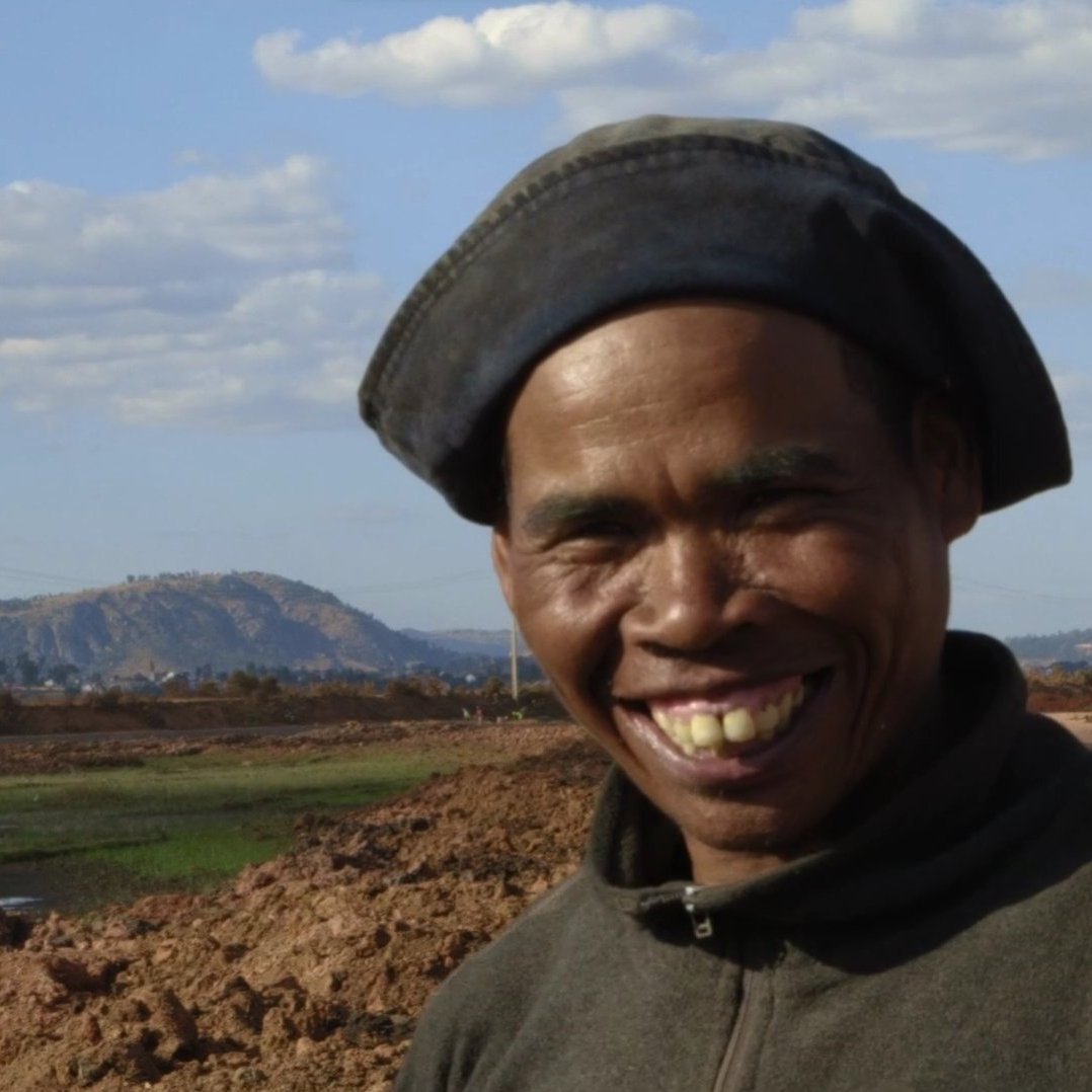 The image features a smiling man wearing a dark hat and a sweater. He is standing in an outdoor setting, likely near agricultural land, with a backdrop of hills and a partly cloudy sky. The ground appears to be bare earth, suggesting possible farming activity nearby. His cheerful expression conveys a sense of warmth and friendliness.