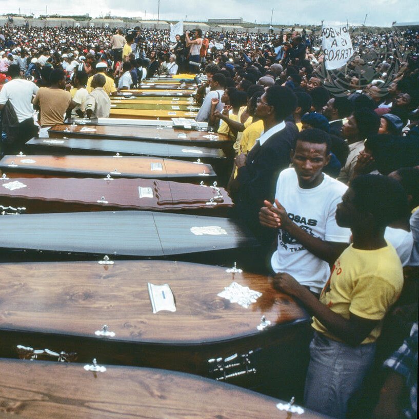 L'image montre une vaste foule rassemblée en extérieur, entourant une série de cercueils alignés. Les cercueils, de différentes couleurs et styles, sont disposés côte à côte, symbolisant une cérémonie funèbre. Les personnes présentes semblent nombreuses et engagées, certaines portent des vêtements de deuil, tandis que d'autres affichent des expressions de tristesse ou de solidarité. L'atmosphère est empreinte de respect et de recueillement, évoquant une perte collective. Au fond, des pancartes sont visibles, ajoutant une dimension politique ou sociale à l'événement. Le ciel est nuageux, renforçant le caractère solennel de la scène.
